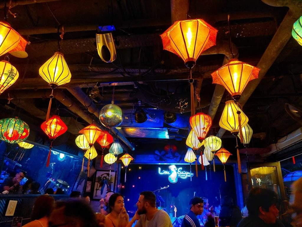 A lively bar atmosphere with colourful lanterns hanging from the ceiling, people socialising, and a live band performance in the background.