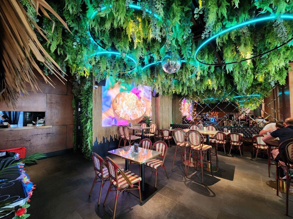 A vibrant bar with lush greenery overhead, neon lights, and a mix of bar stools and tables, with a large screen displaying visuals.