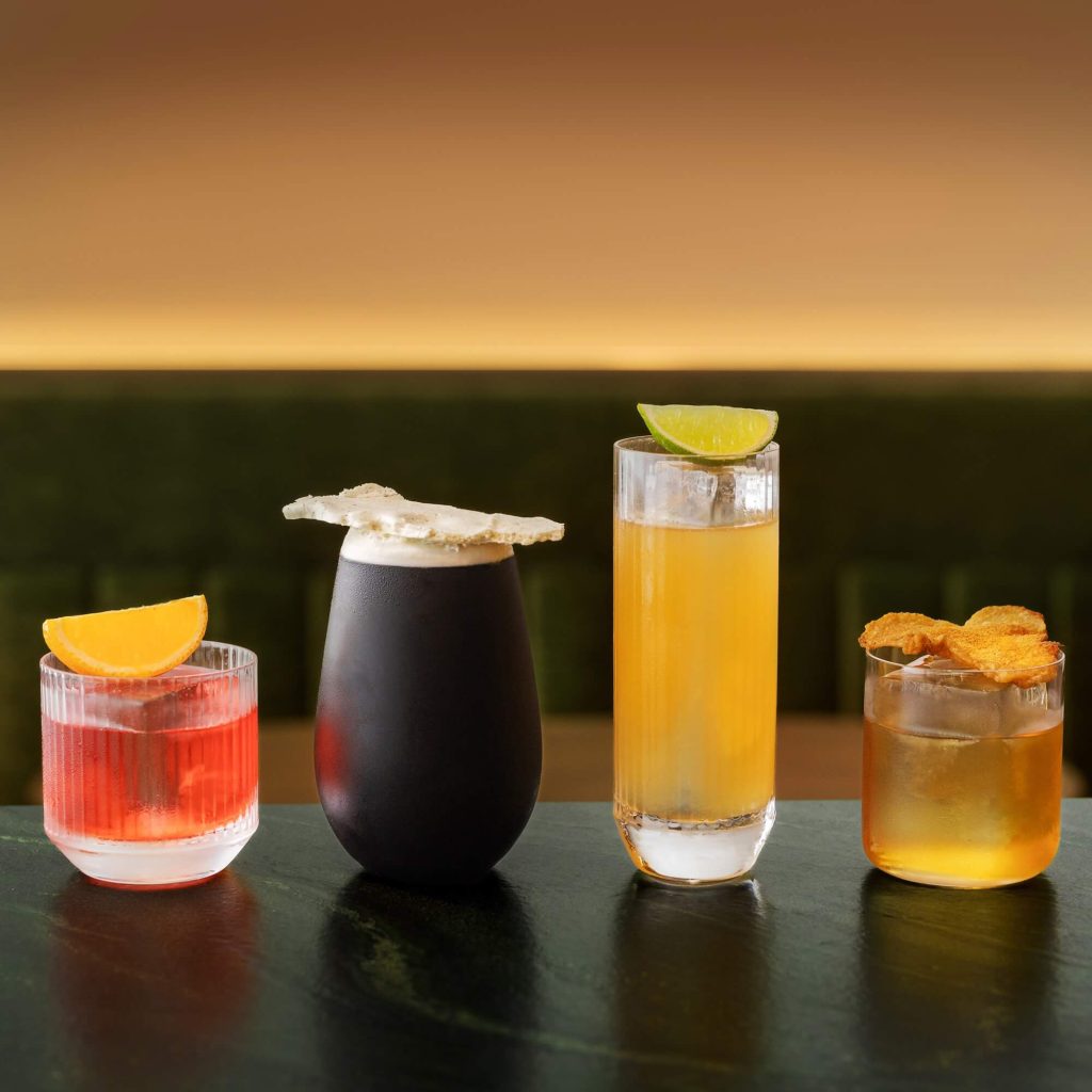 Four cocktails on a bar, each with a unique garnish, against a warm, gradient backdrop, offering a variety of colours and glassware.
