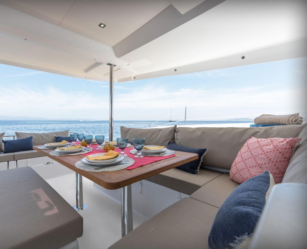 A dining setup on a yacht with a wooden table set for a meal, comfortable seating, and a view of the sea and distant coastline.