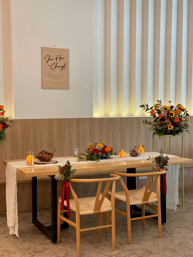 Cafe table with a floral setup and lit candles, with two wooden rattan chairs and a bench vis a vis.