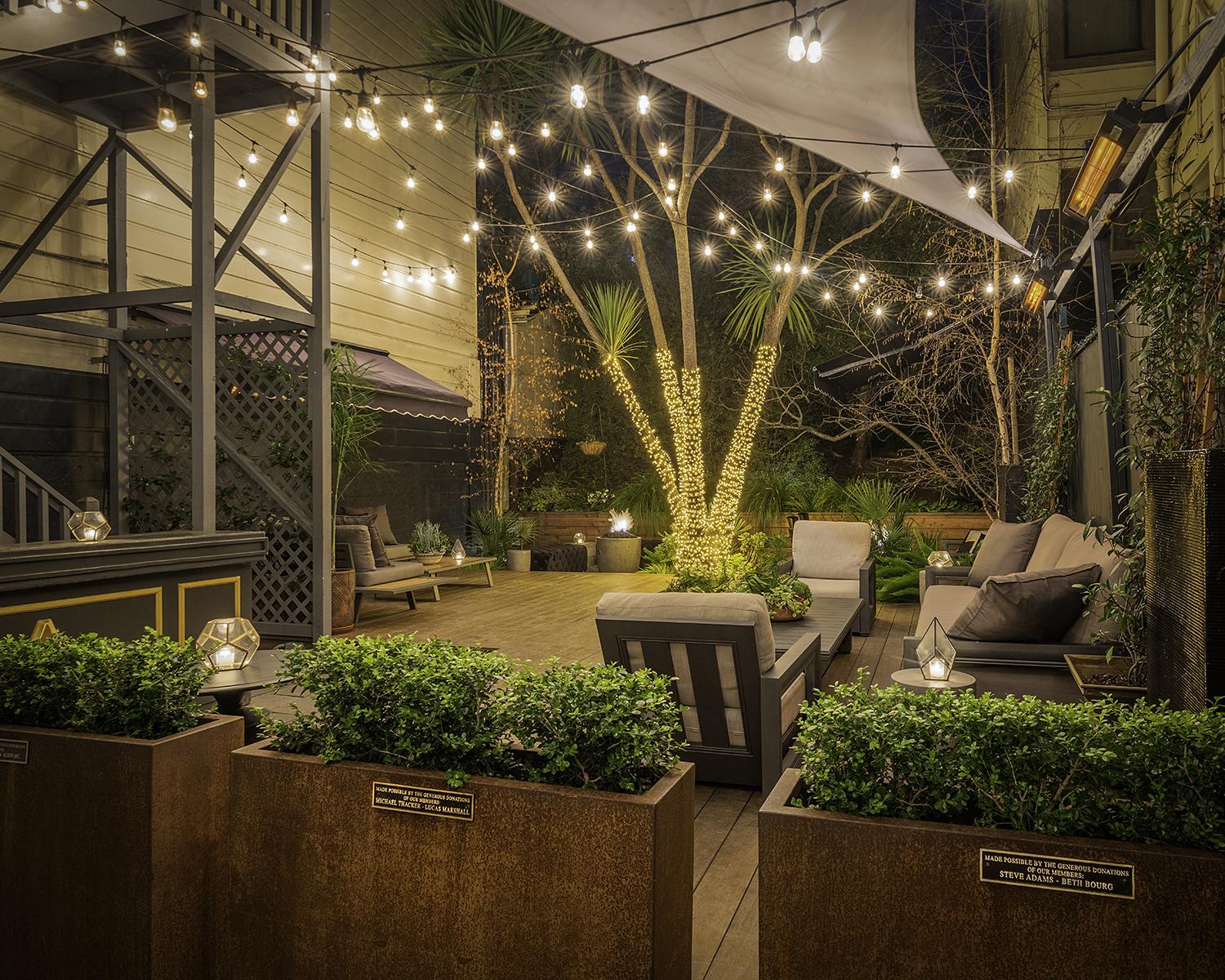 An outdoor bar area with string lights, lounge seating, and a tree wrapped in lights at The Academy.