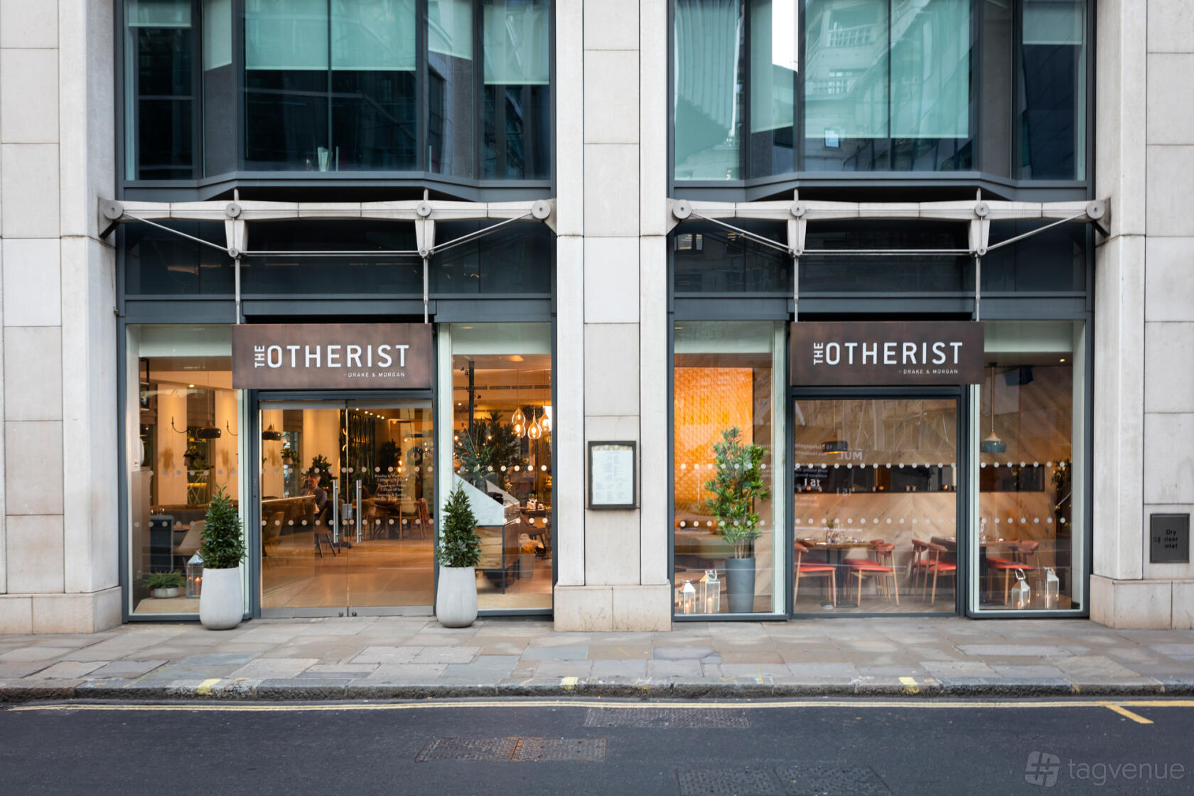 A restaurant exterior with large glass windows, potted plants, and warm interior lighting at The Otherist.