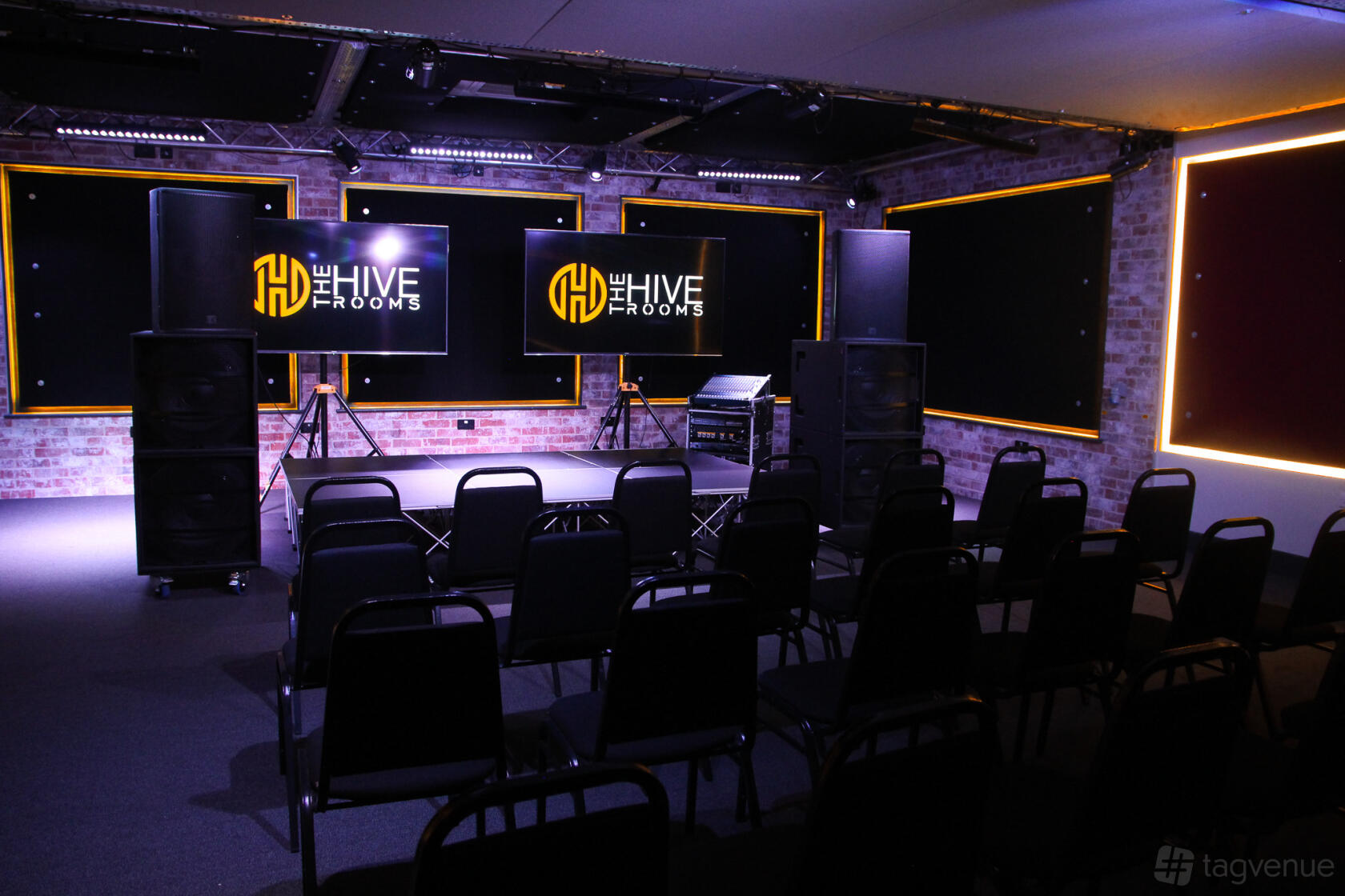 A recording studio with exposed brick walls, stage lighting, and chairs arranged facing three large screens at The Hive Rooms.