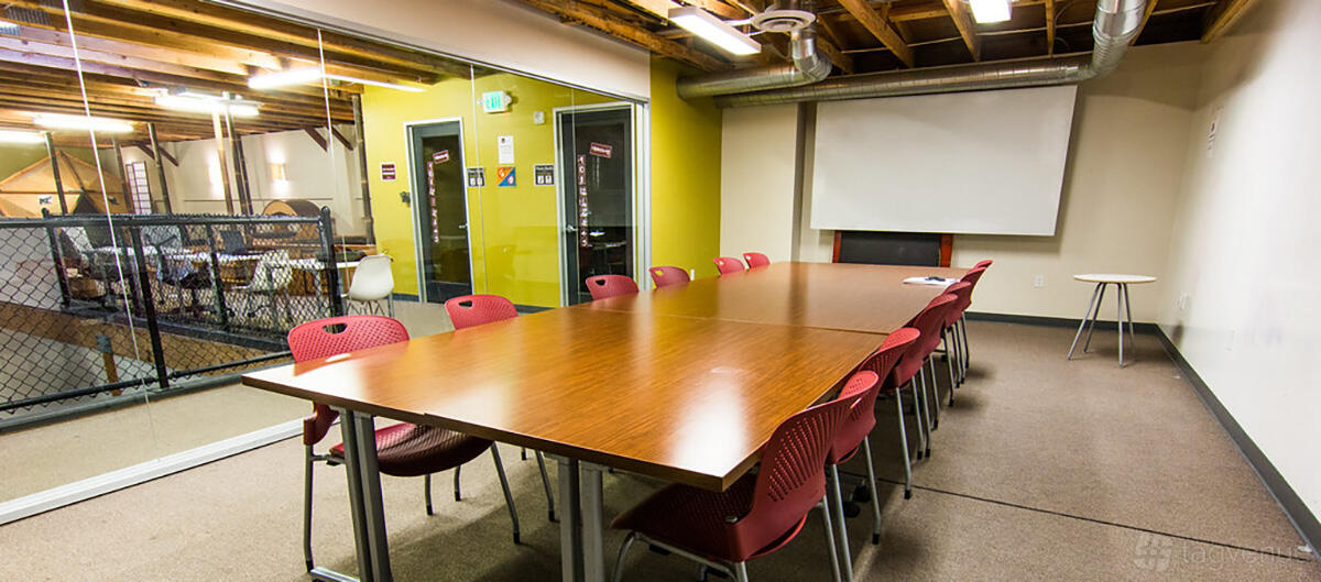 A meeting room with a long rectangular table, red chairs, exposed ceiling beams, and a projector screen at Oakstop 2323 Broadway.
