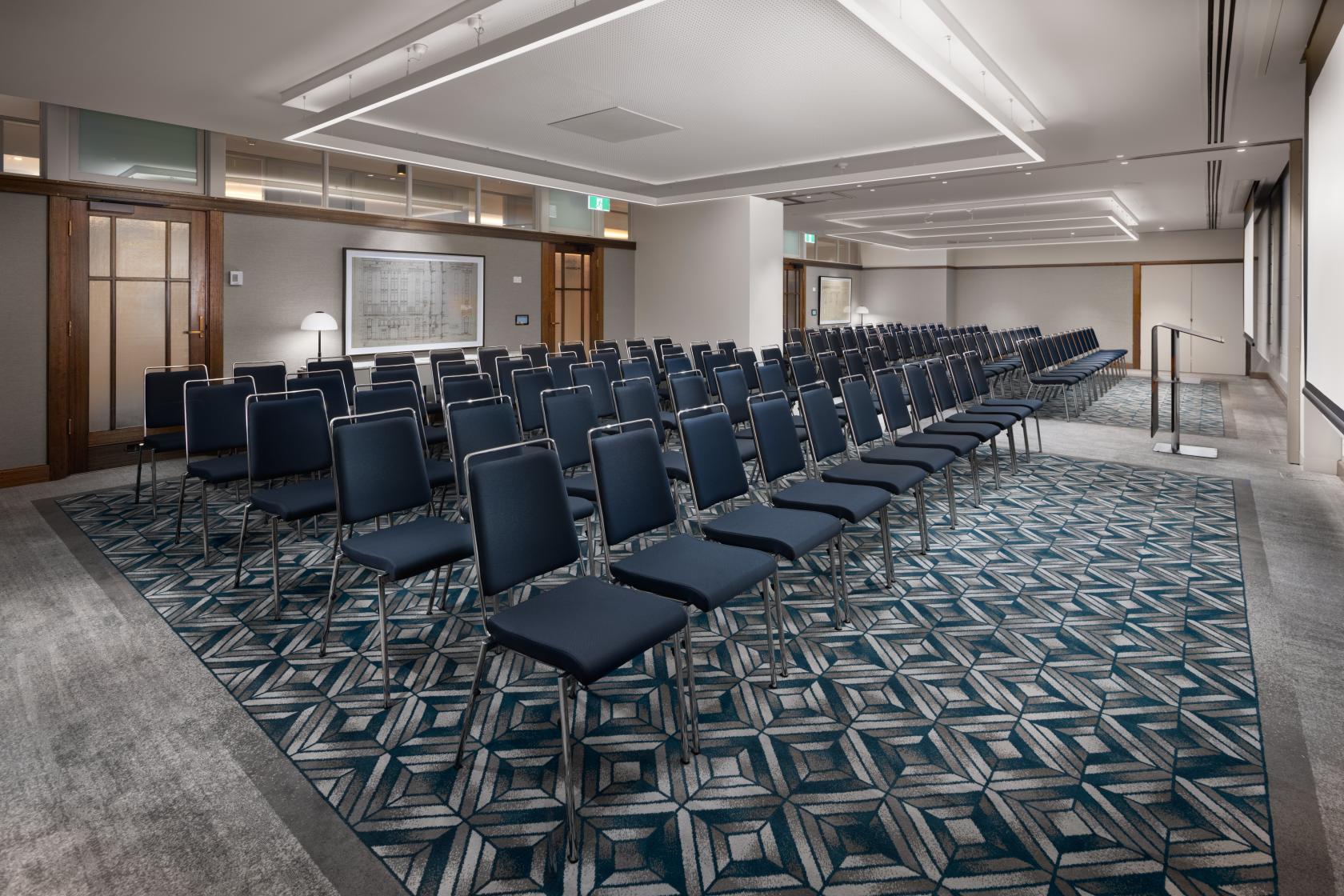 An auditorium with rows of navy chairs, geometric carpet, and floor-to-ceiling windows at Hilton Melbourne Little Queen Street.