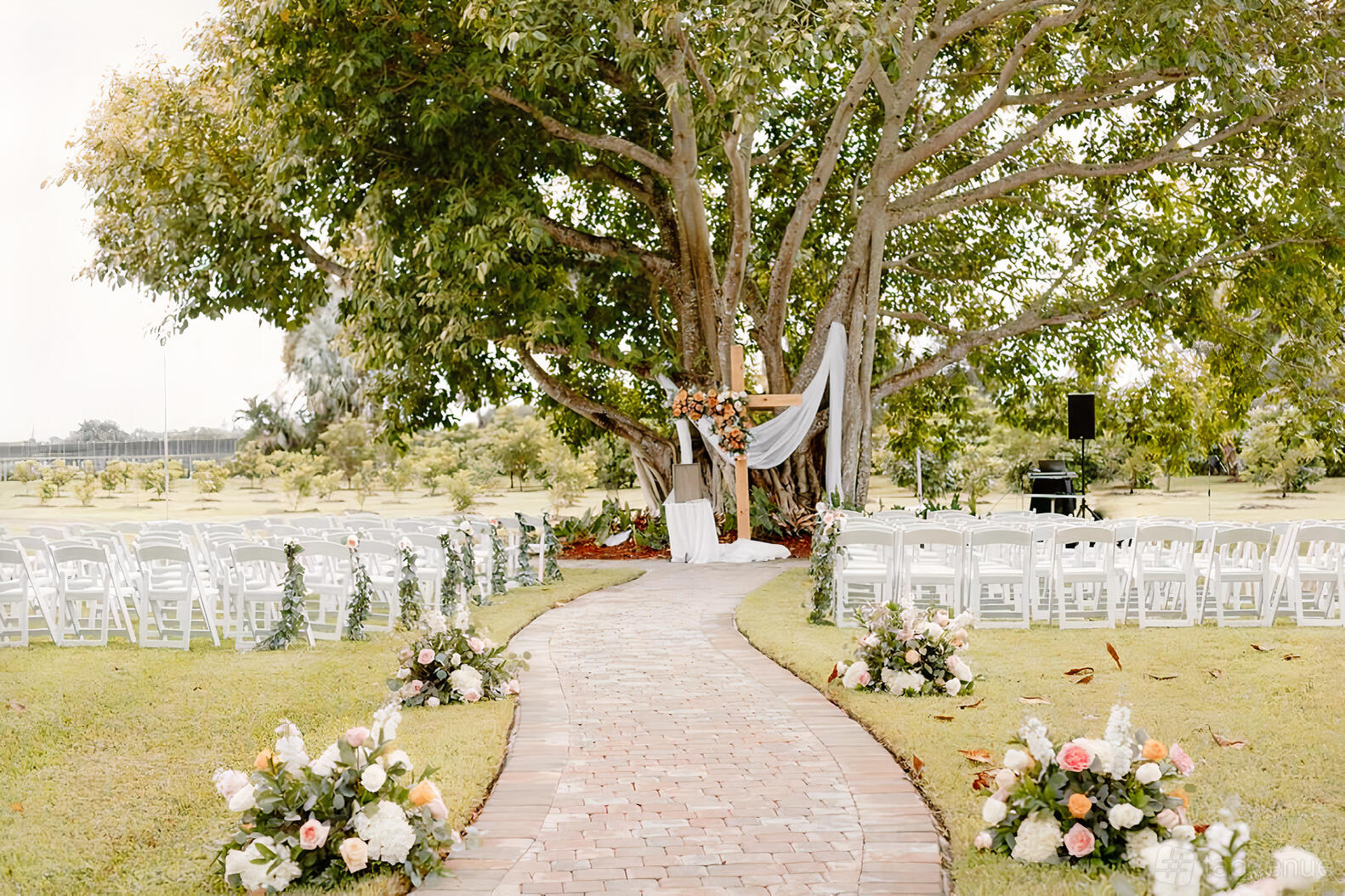 An outdoor event venue with a brick aisle, white chairs, floral arrangements, and a large tree altar at Cinco Farm.