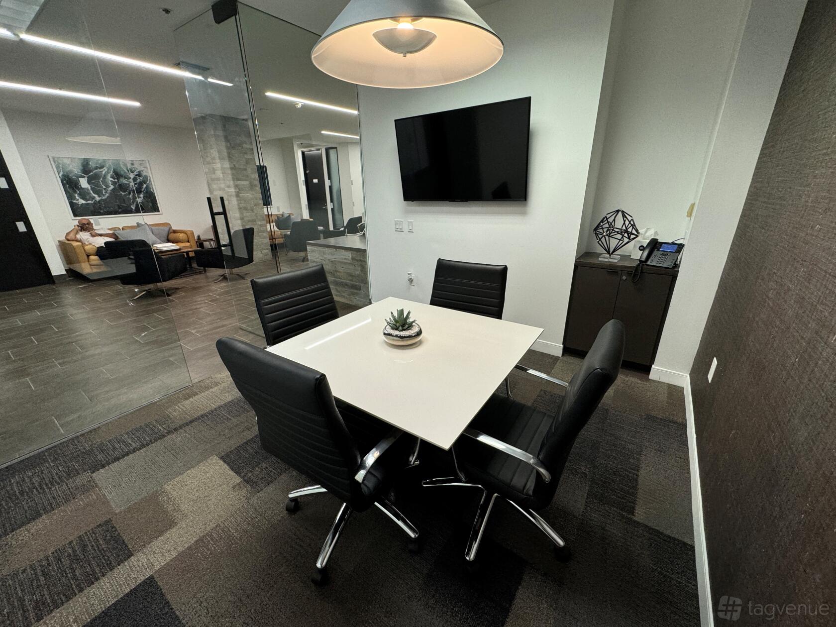 A coworking boardroom with a square table, four black chairs, wall-mounted screen, and glass wall at Premier Workspaces - Huntington Beach.