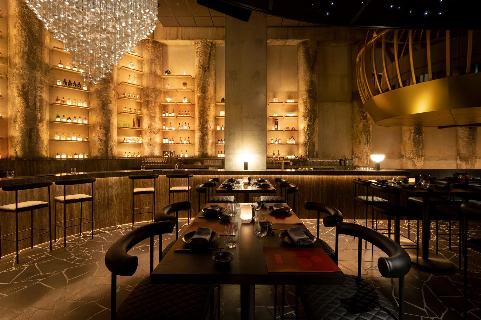 A restaurant with backlit shelves of bottles, elegant stone flooring, and a crystal chandelier at Yugen Dining.