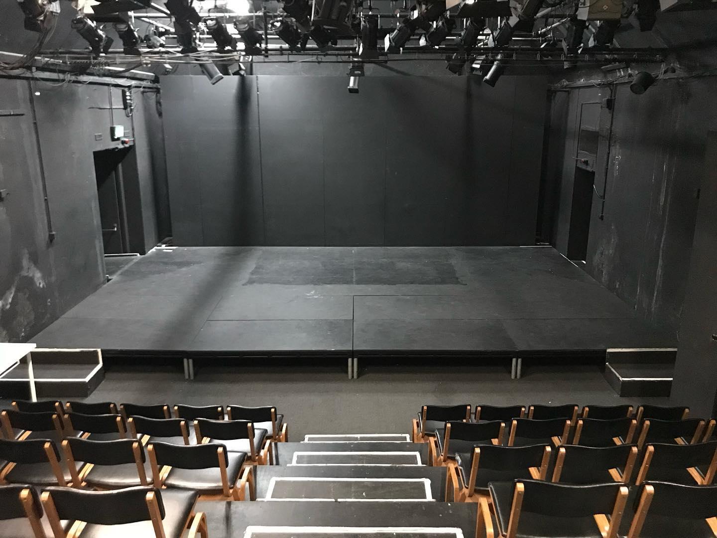 A black box theatre with tiered seating, a raised stage, and overhead lighting rigs at Network Theatre.
