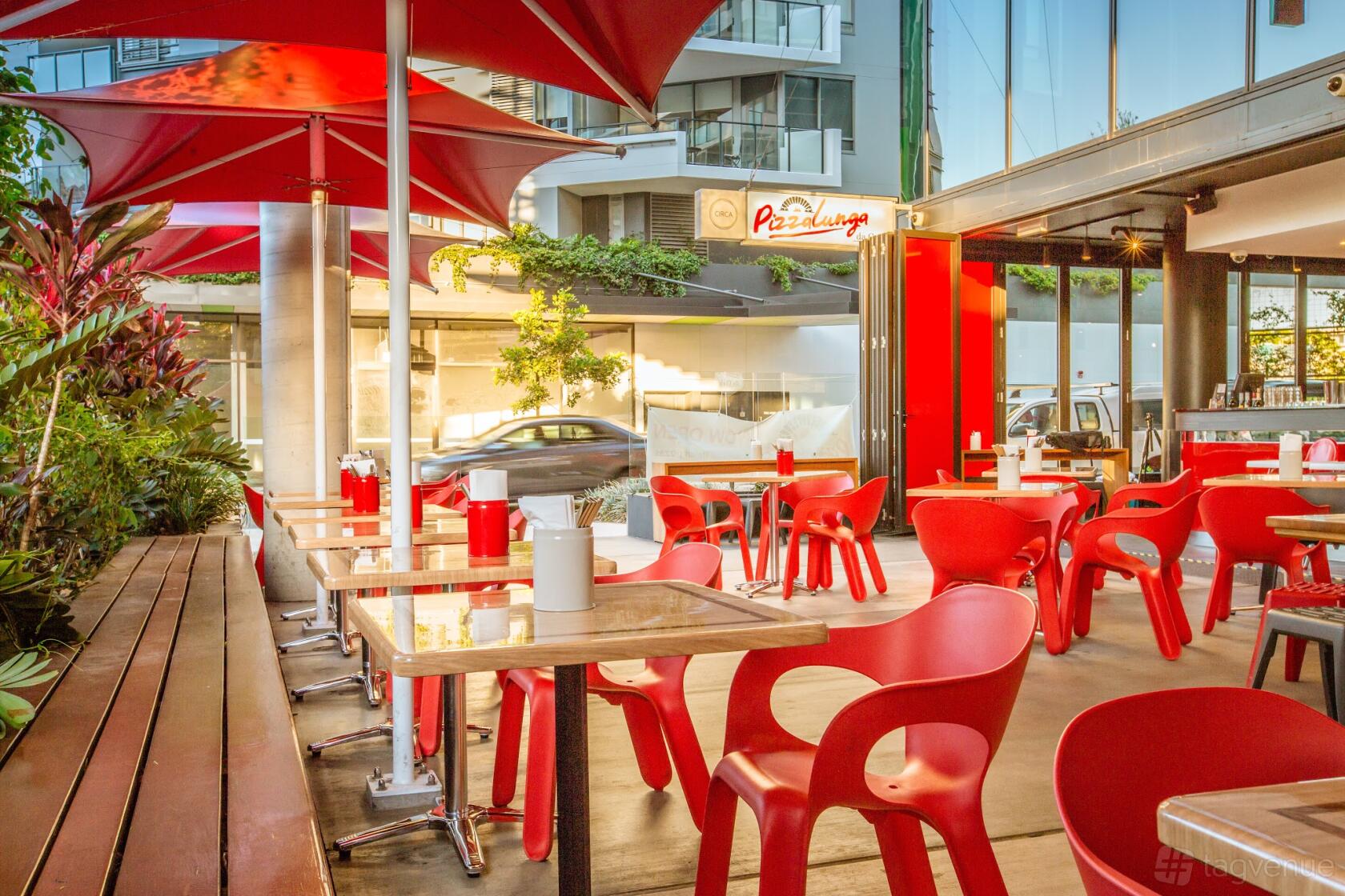 An outdoor restaurant event space with red chairs, wooden tables, and red umbrellas at Pizzalunga da Carlo.