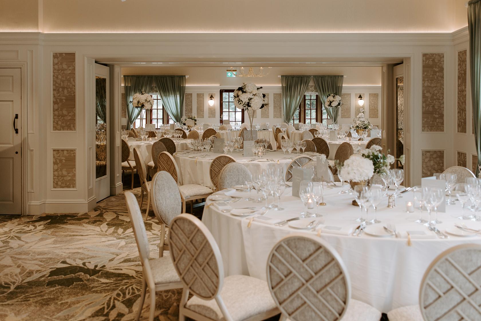 An indoor banquet hall with round tables, floral centerpieces, and upholstered chairs at Pennyhill Park.