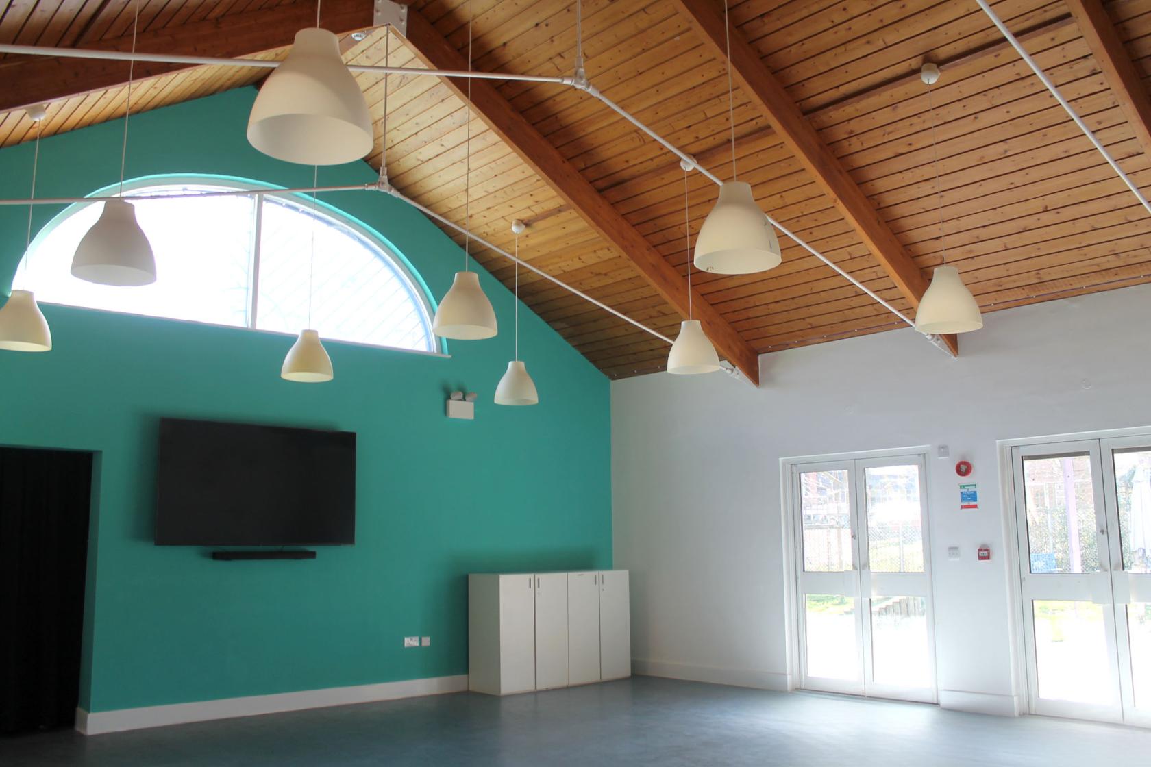 A community hall with vaulted wooden ceilings, hanging pendant lights, and a teal accent wall at Castlehaven Community Centre & Hub.