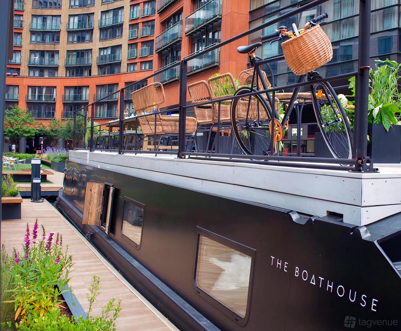 A canal boat with an upper deck featuring rattan chairs, a bicycle, and potted plants at The Boathouse London - Paddington West.