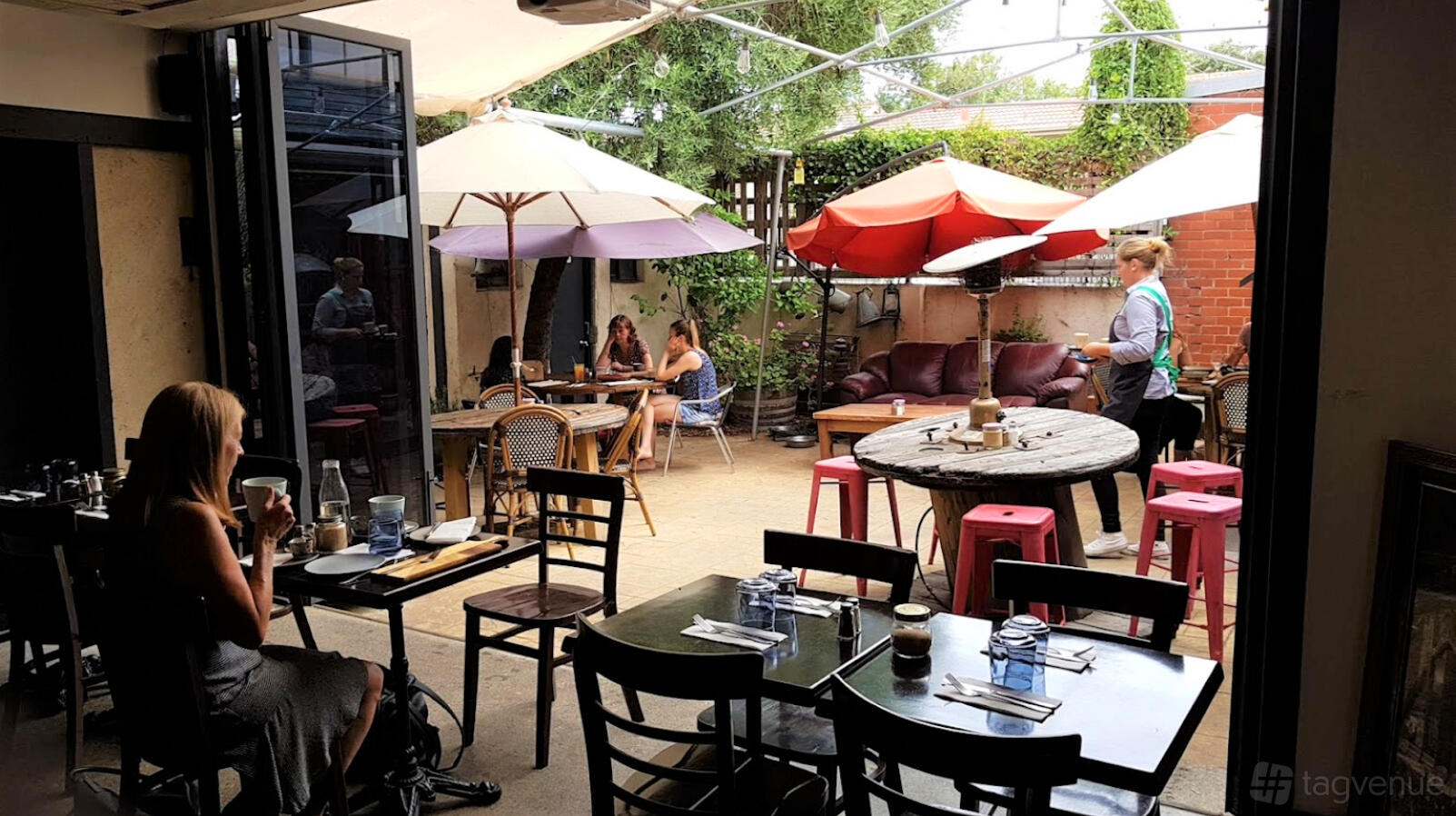 An outdoor restaurant patio with round tables, mix of chairs and stools, and colorful umbrellas at The Little French Deli & Bistro.