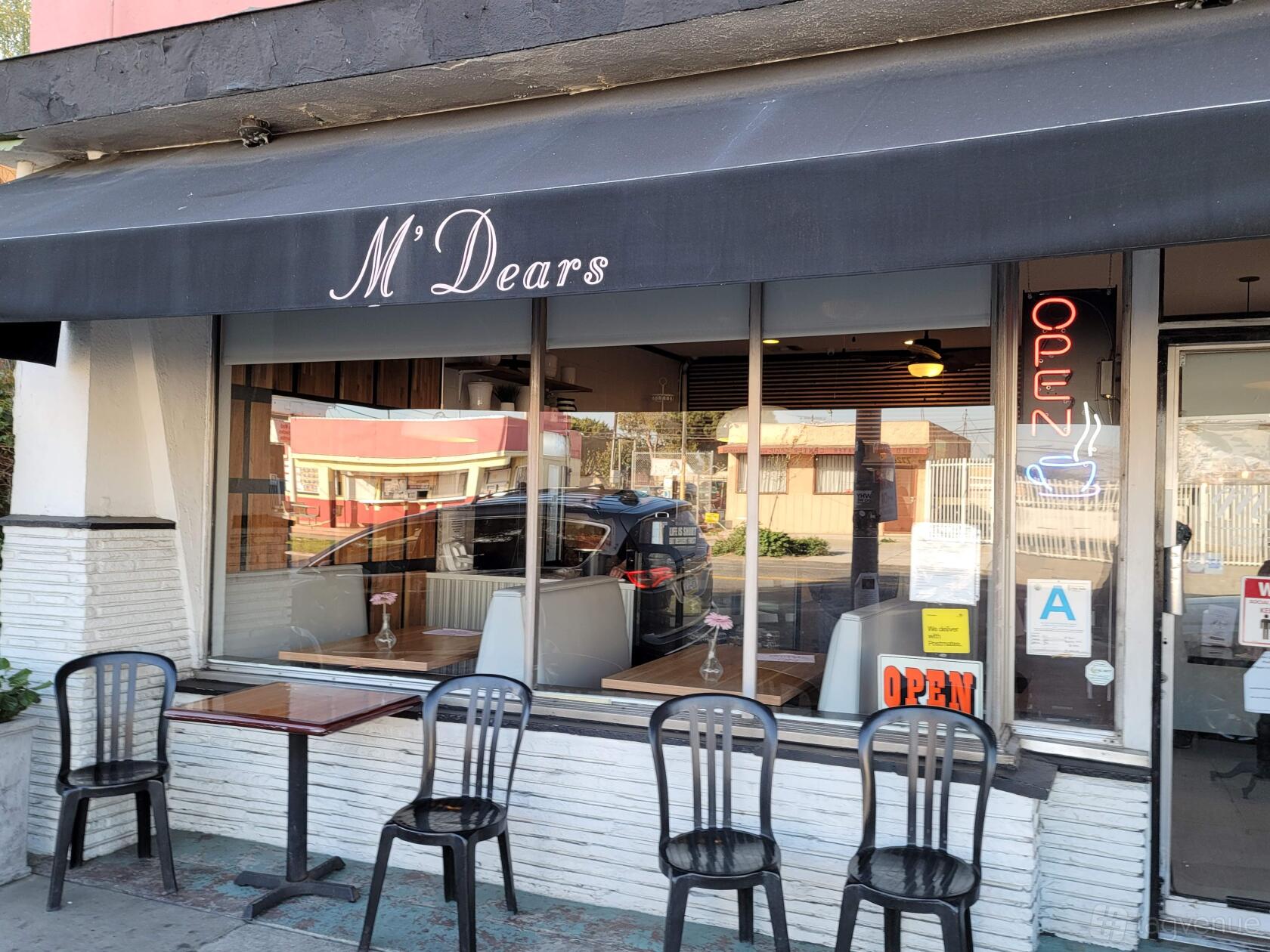 A restaurant with sidewalk seating, black chairs, and a large window under a black awning at M'Dears Bakery & Bistro.