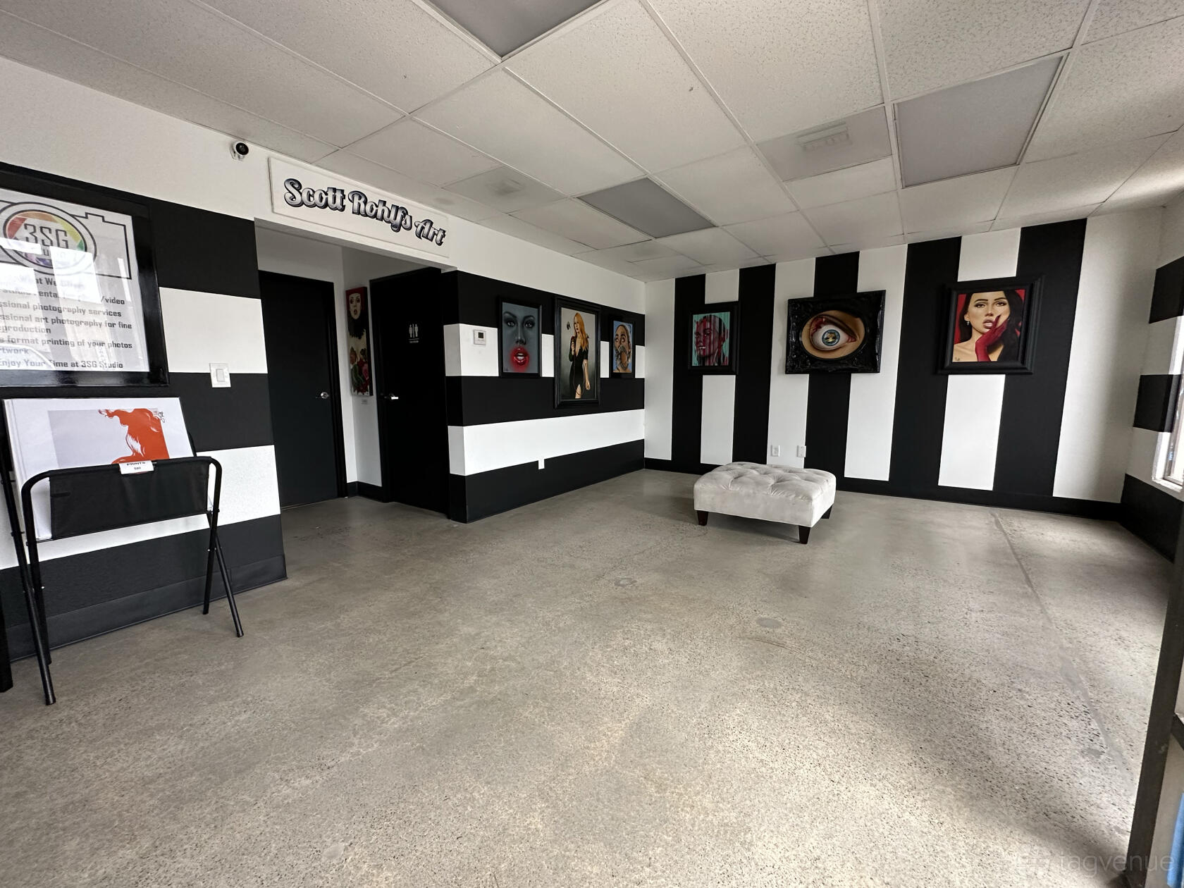 A photo studio with black and white striped walls, concrete floors, portrait art, and a central ottoman at 3SG Studio.