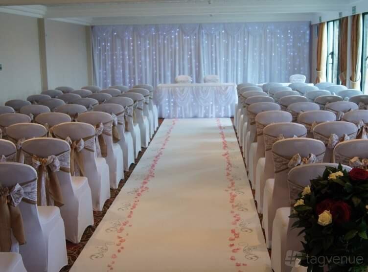 An event space in a hotel with rows of white chair covers and a petal-lined aisle at Hellidon Lakes Golf & Spa Hotel.