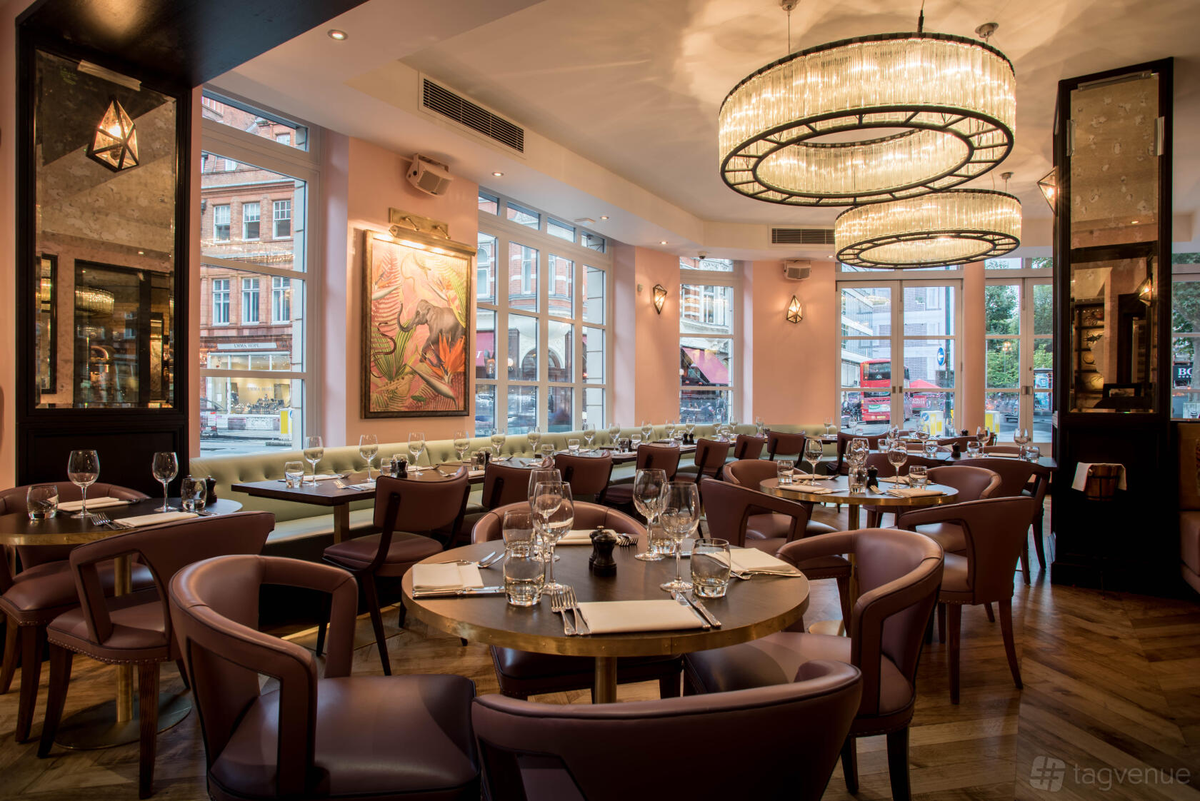 A dining room in a cocktail bar with large windows, round tables, and two circular chandeliers at The Botanist Sloane Square.