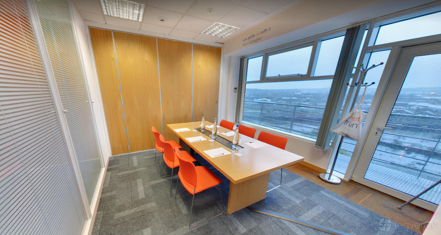 A meeting room with floor-to-ceiling windows, orange chairs, and a wooden table at The Studio Leeds.
