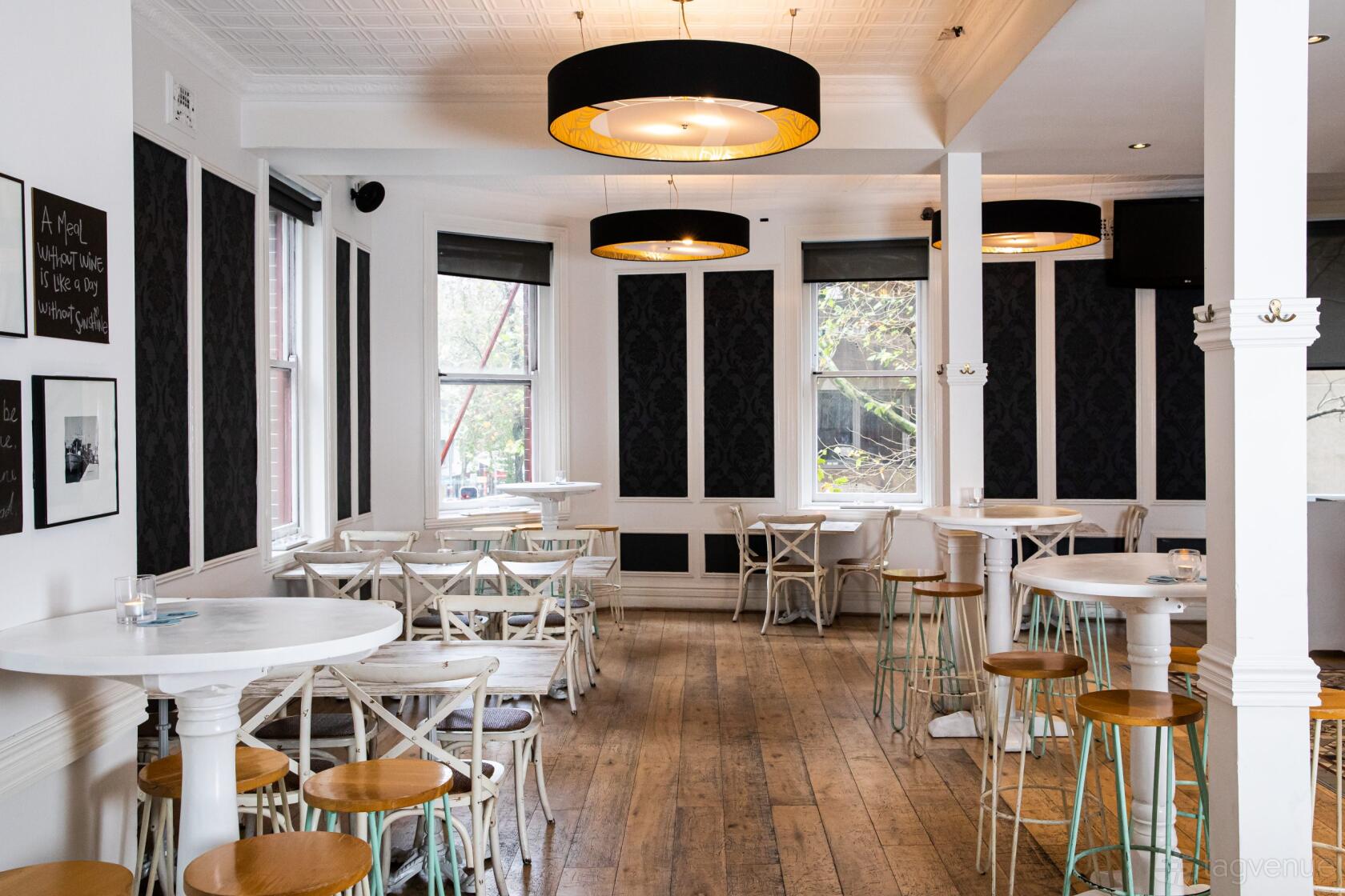 An event space in a pub with high ceilings, round white tables, wooden stools, and large windows at The Belvedere Hotel.
