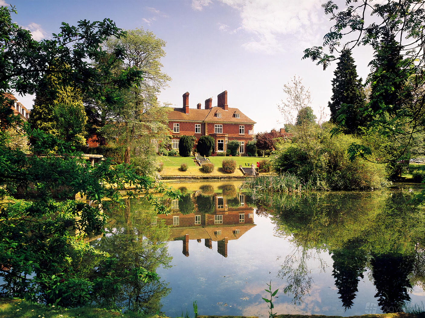 A historic hotel with red brick exterior, tall chimneys, and landscaped gardens overlooking a pond at Albrighton Hall Hotel.