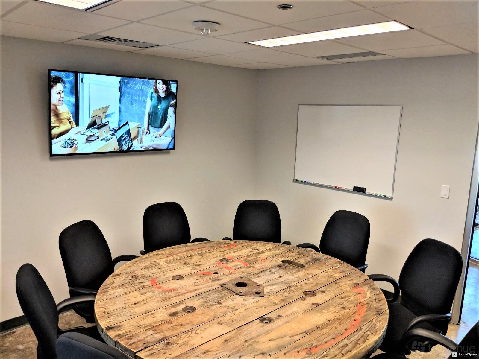 A boardroom with a round wooden table, black office chairs, wall-mounted screen, and whiteboard at Vessel Coworking.