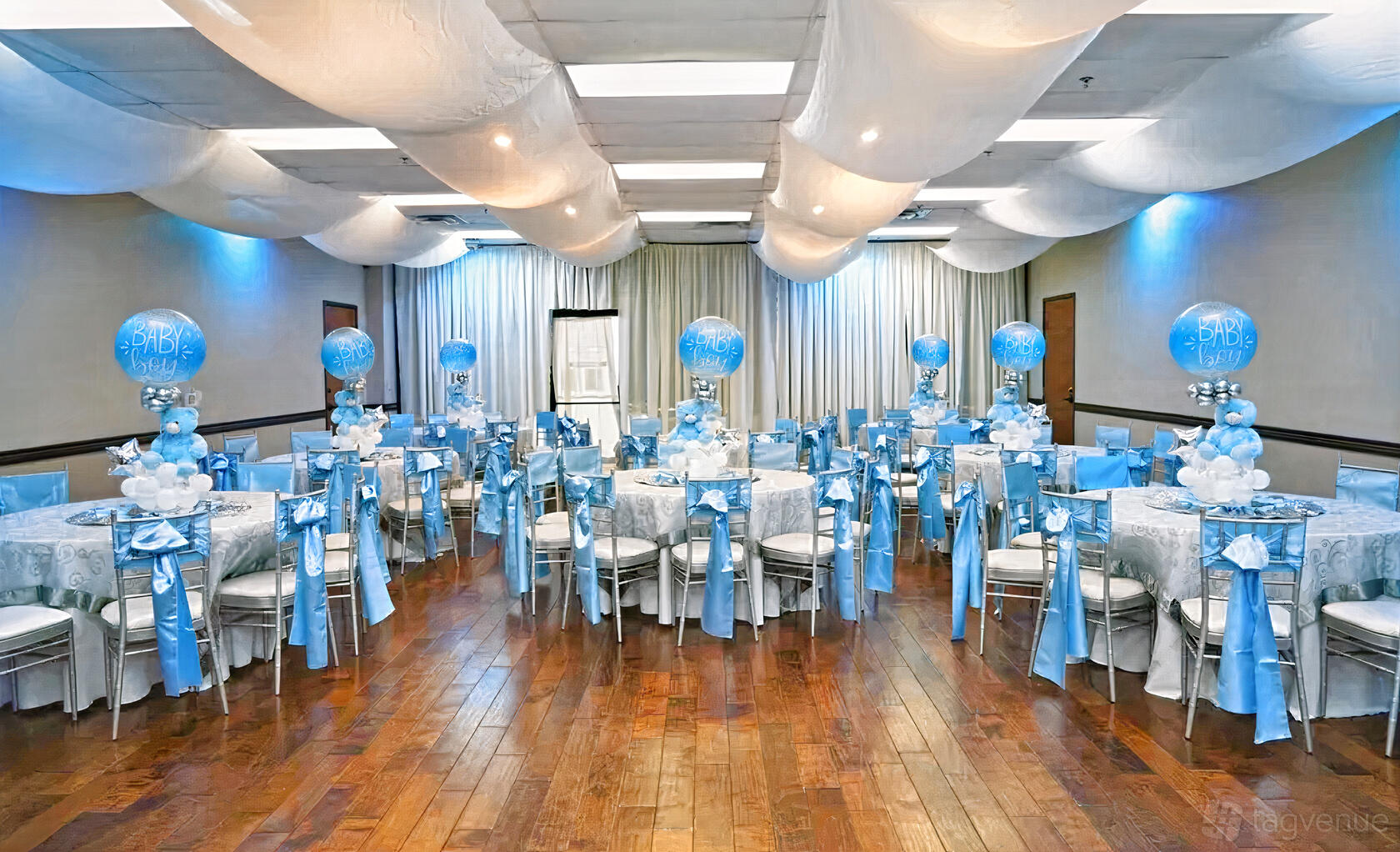 A banquet hall with polished wood floors, white draped ceiling, and round tables decorated with blue balloons at The Oasis Ballroom.