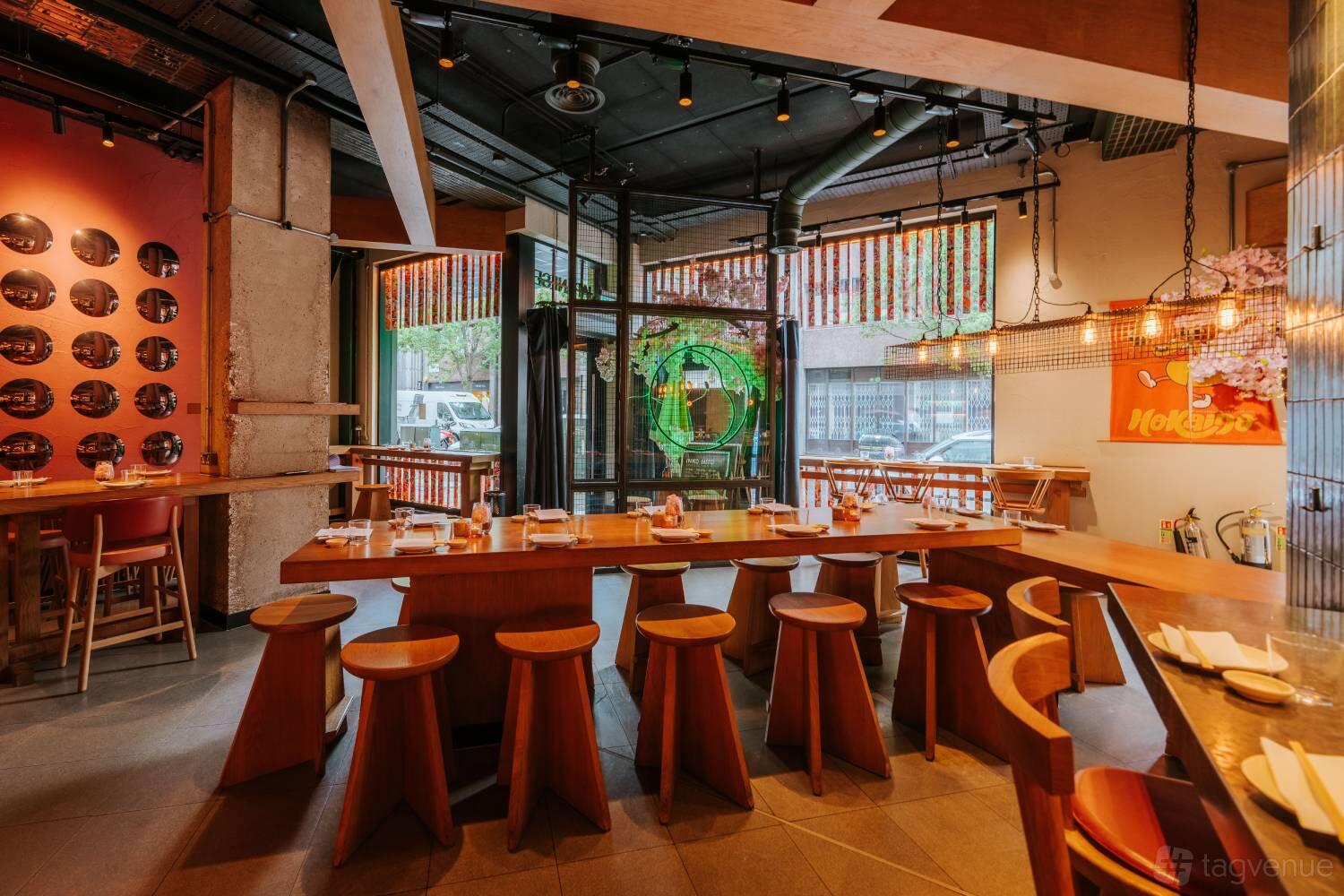 A restaurant with wooden communal tables, round stools, and decorative hanging lights at INKO NITO London.