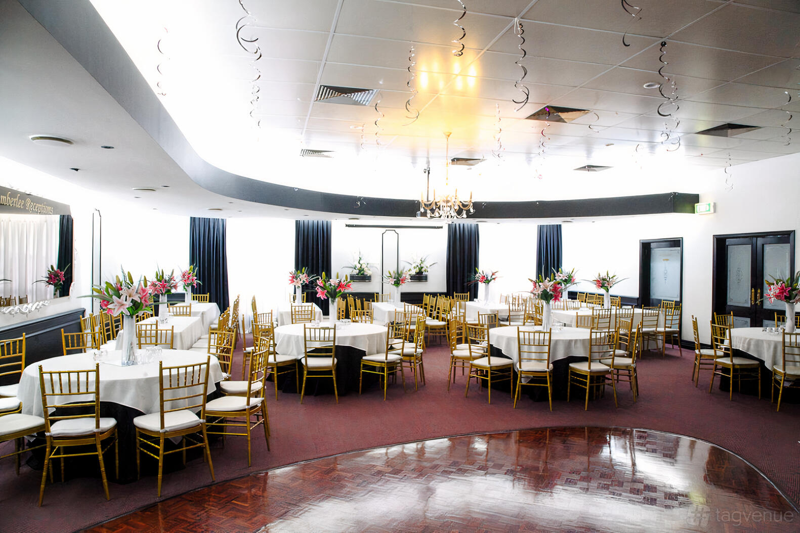 A ballroom with round tables, gold chairs, floral centerpieces, and a parquet dance floor at Amberlee Receptions.