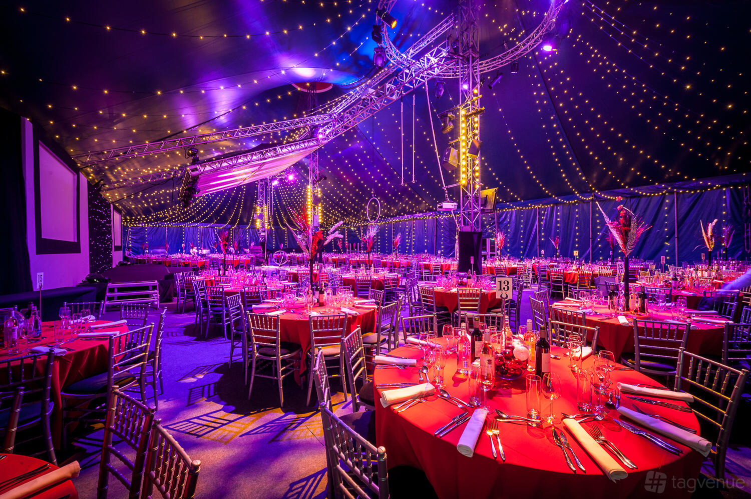 An event venue with round tables draped in red linens, chiavari chairs, and ceiling string lights at Bloomsbury Big Top.