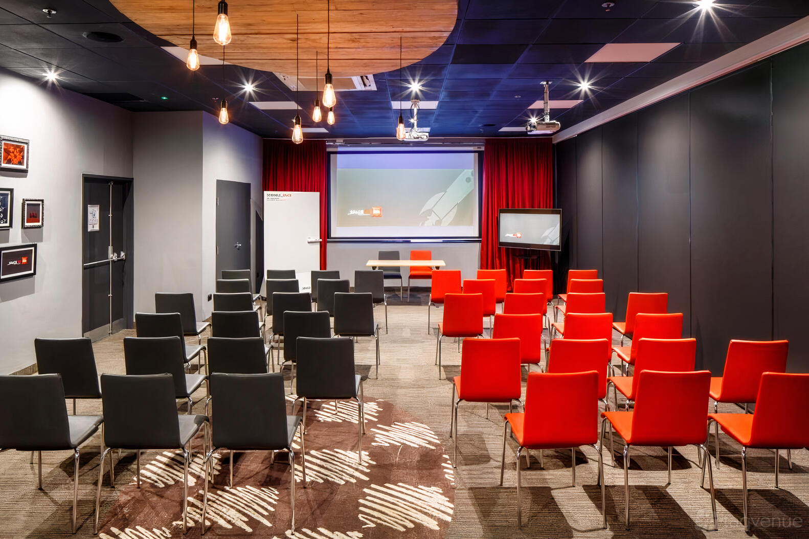 An event space with rows of black and red chairs, stage, projector screen, and pendant lights at Ibis Birmingham New Street.