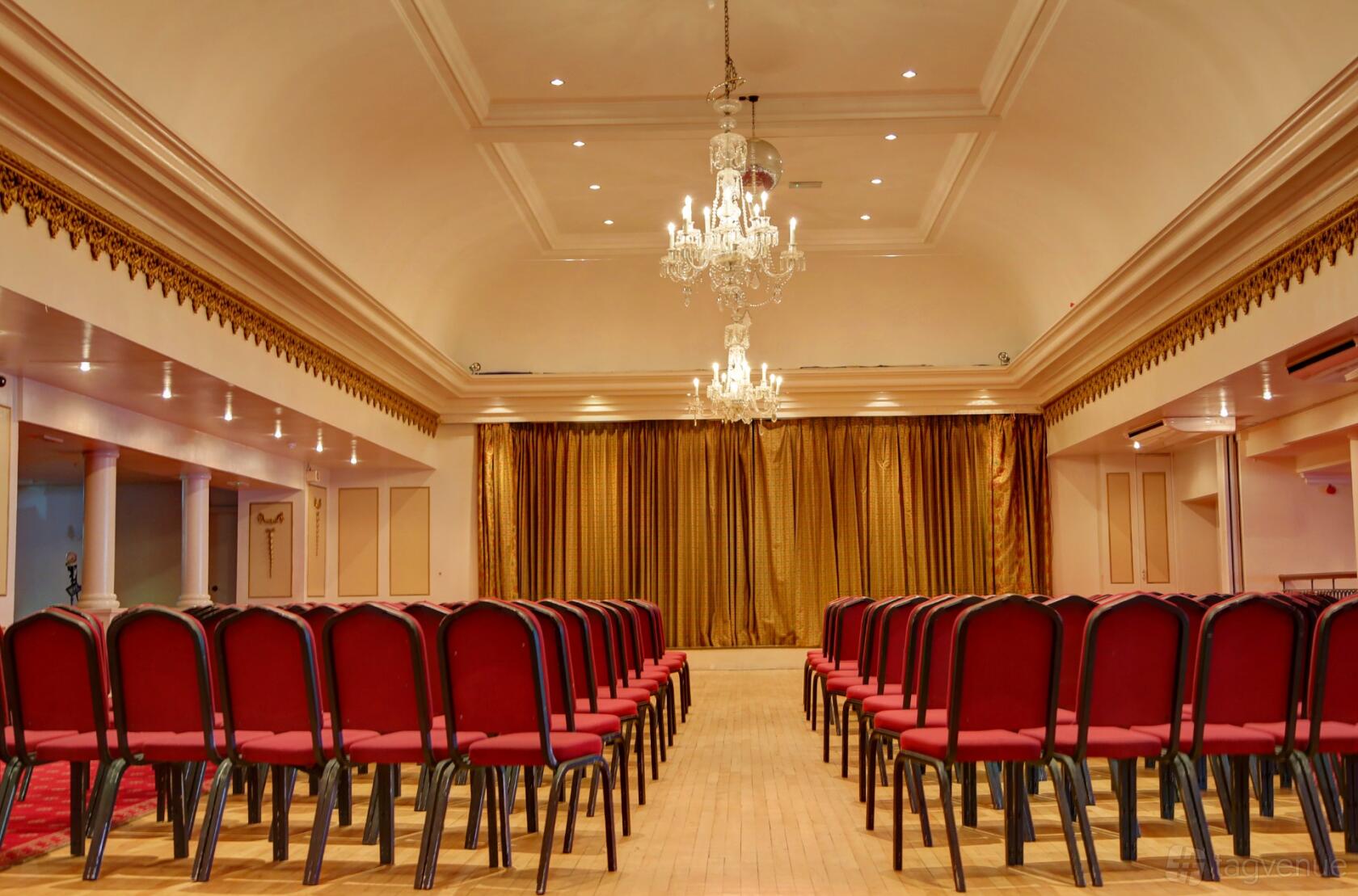 A hotel ballroom with red upholstered chairs, chandeliers, and gold stage curtains at The Craiglands Hotel.