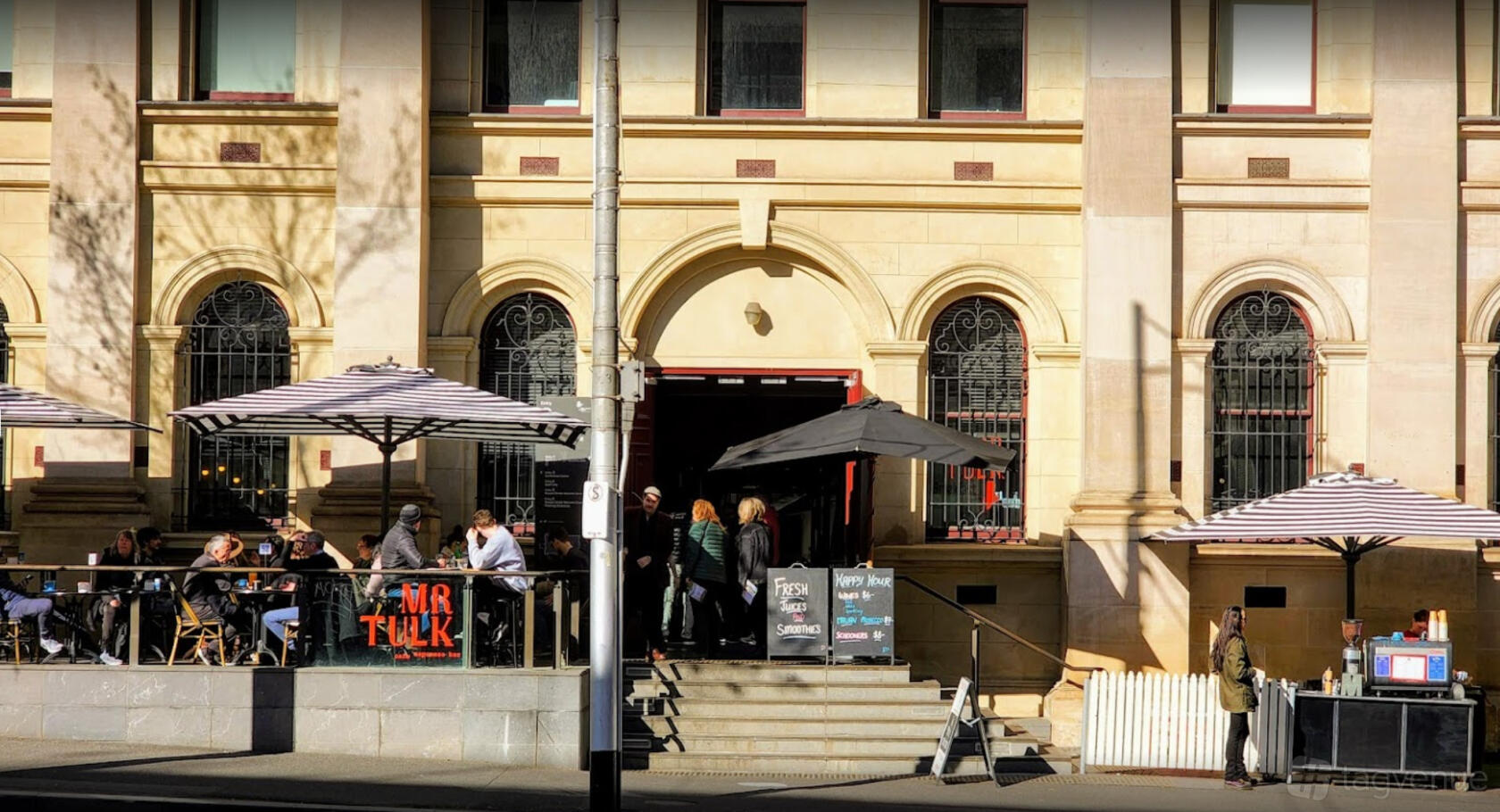 A café with striped patio umbrellas, outdoor seating, and arched windows at Mr Tulk Cafe.