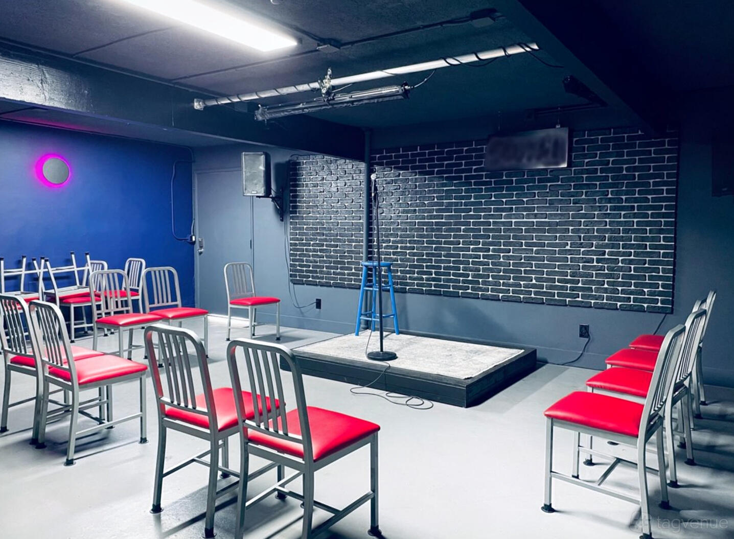 An event venue with a small stage, exposed brick wall backdrop, red-seated chairs, and a blue accent wall at The Nitecap.