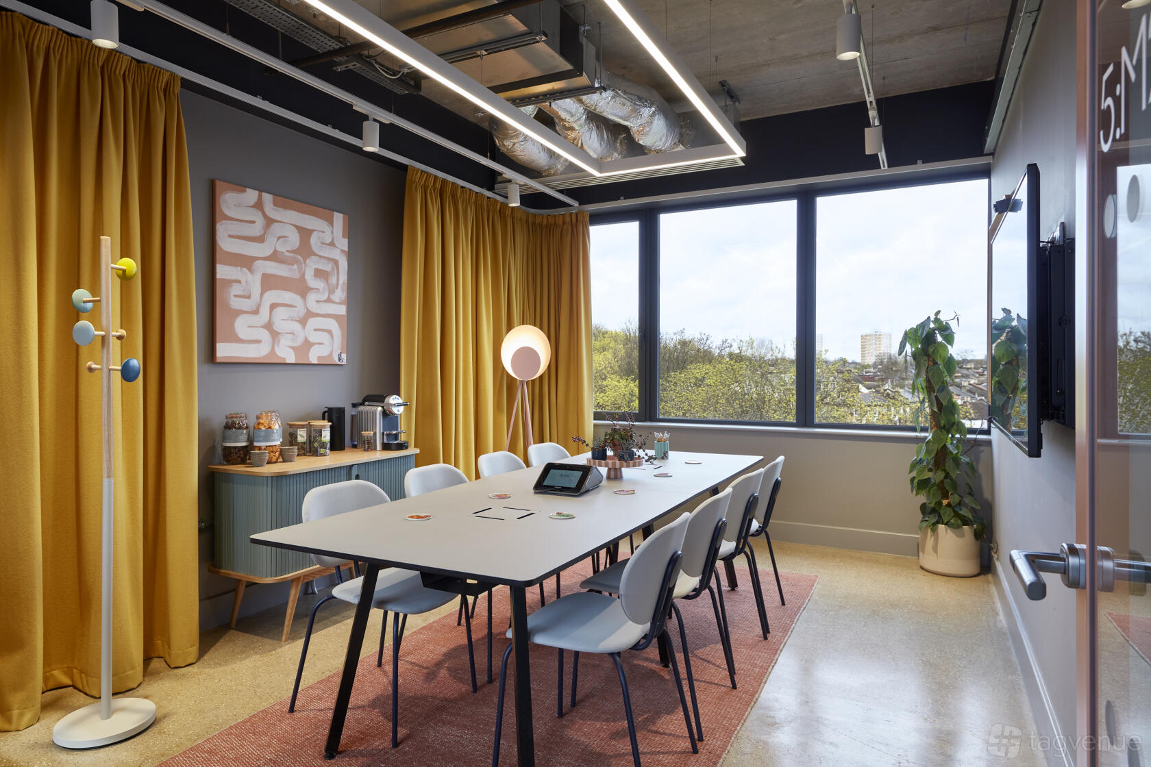A coworking space with large windows, a long table with eight chairs, mustard curtains, and indoor plants at Clockwise Offices Wood Green.