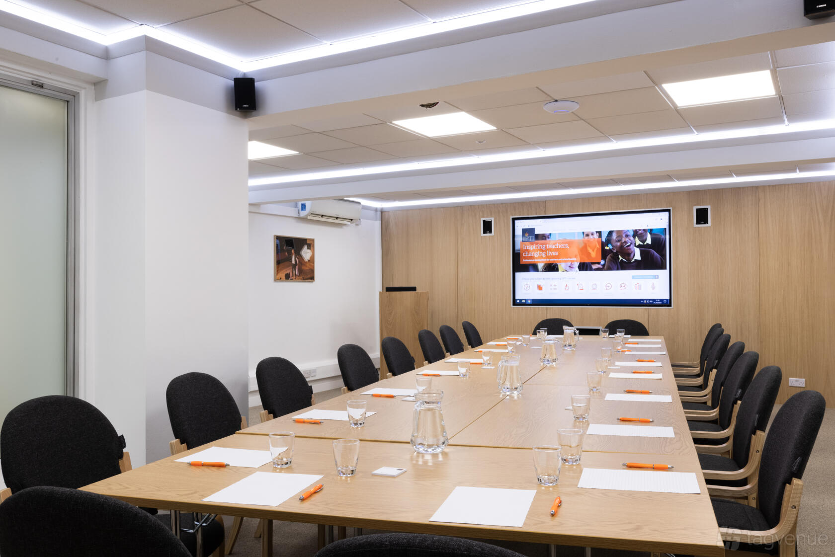 A meeting centre with a long rectangular table, black chairs, wood paneling, and a large digital screen at The Hintze Rooms.