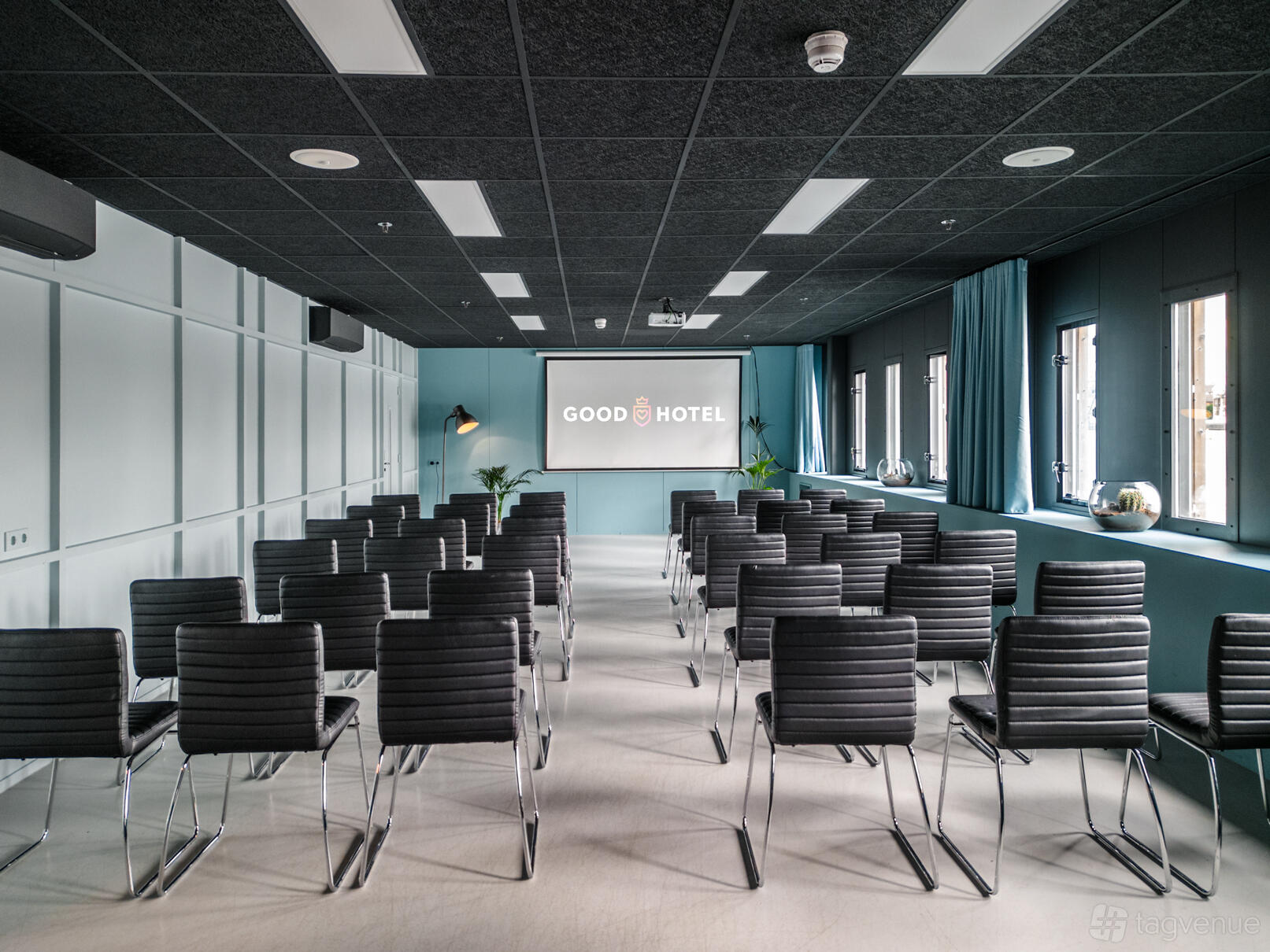 An event space with rows of black chairs, floor-to-ceiling windows, and a projector screen at Good Hotel London.