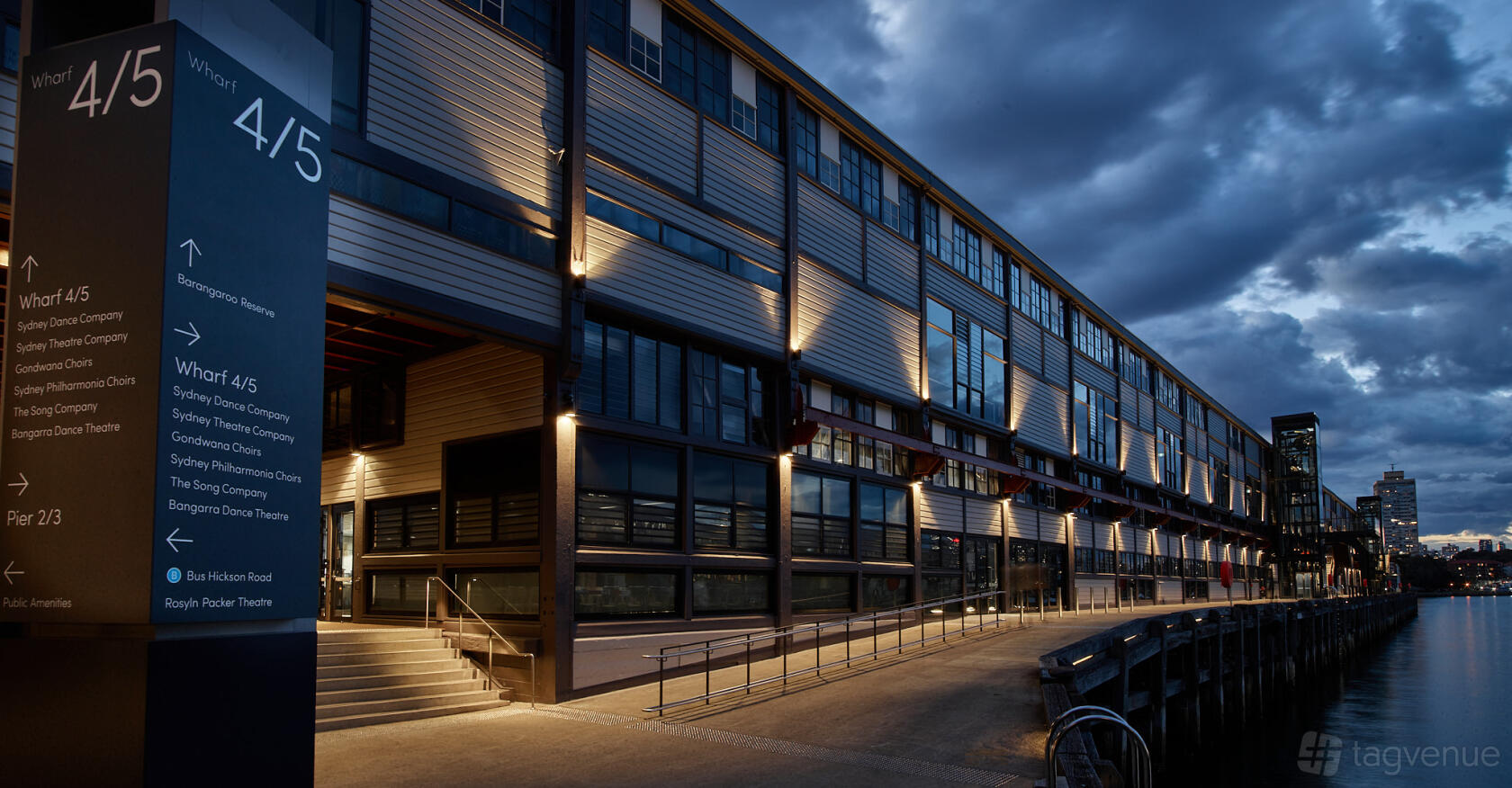 An exterior view of a warehouse-style dance studio with large windows and waterfront walkway at Sydney Dance Company.