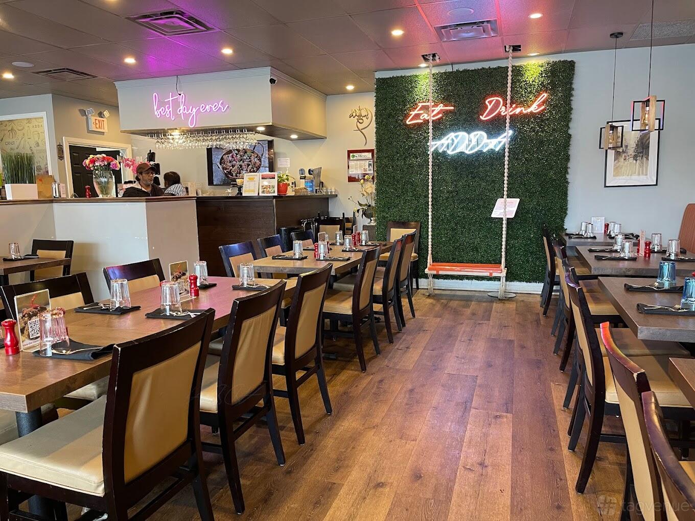 An event space in a restaurant with wood floors, rows of tables with place settings, and a neon sign on a green wall at ADDA Bengali Kitchen & Catering.