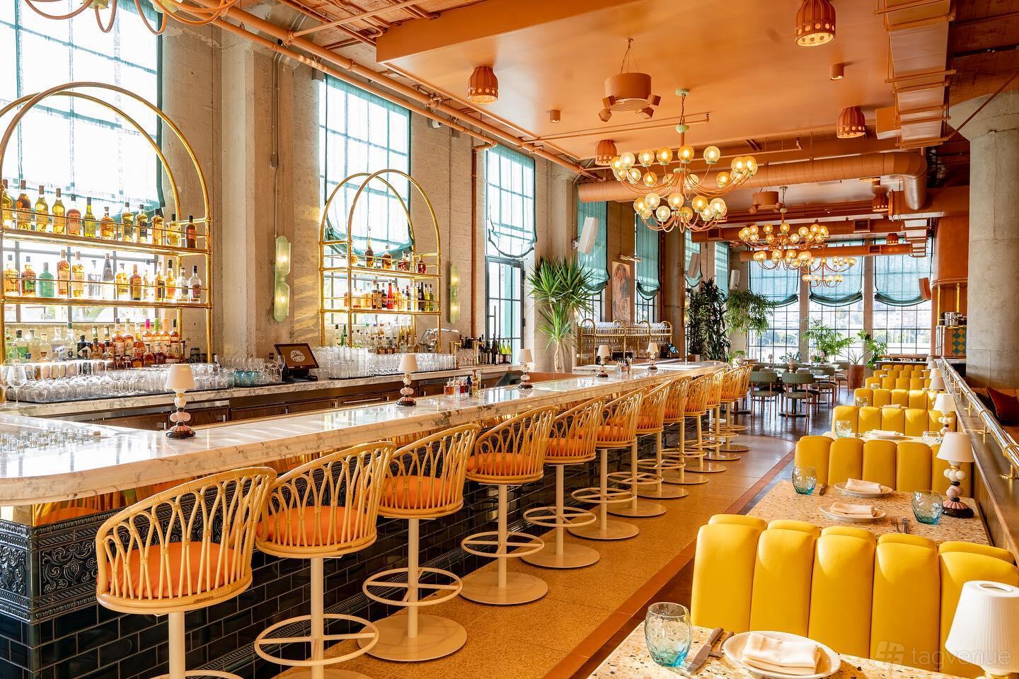 A dining room with a marble-topped bar, yellow upholstered chairs, globe pendant lighting, and large windows at Mother Tongue Restaurant.