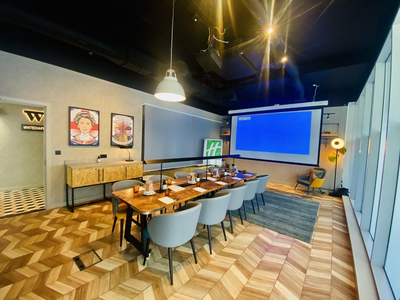 A meeting room with a long wooden table, grey chairs, a projector screen, and framed wall art at Holiday Inn London Whitechapel.