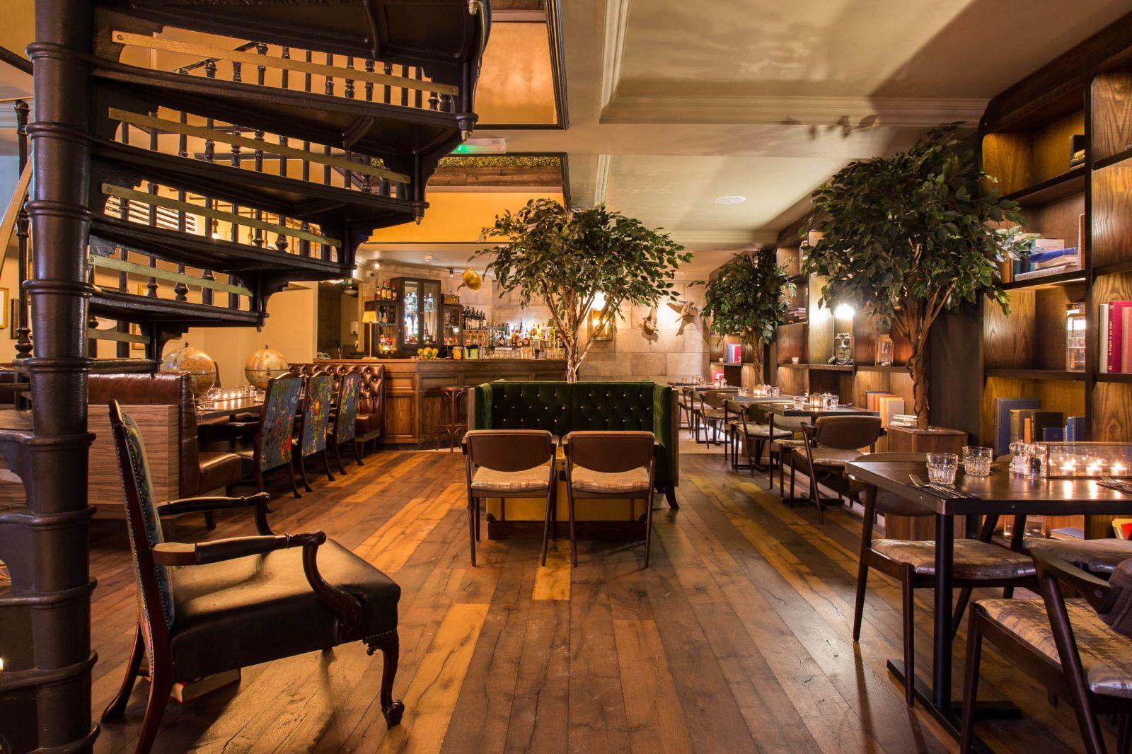 A function room in a bar with wooden floors, spiral staircase, bookshelves, and indoor trees at The Lost and Found Greek Street.