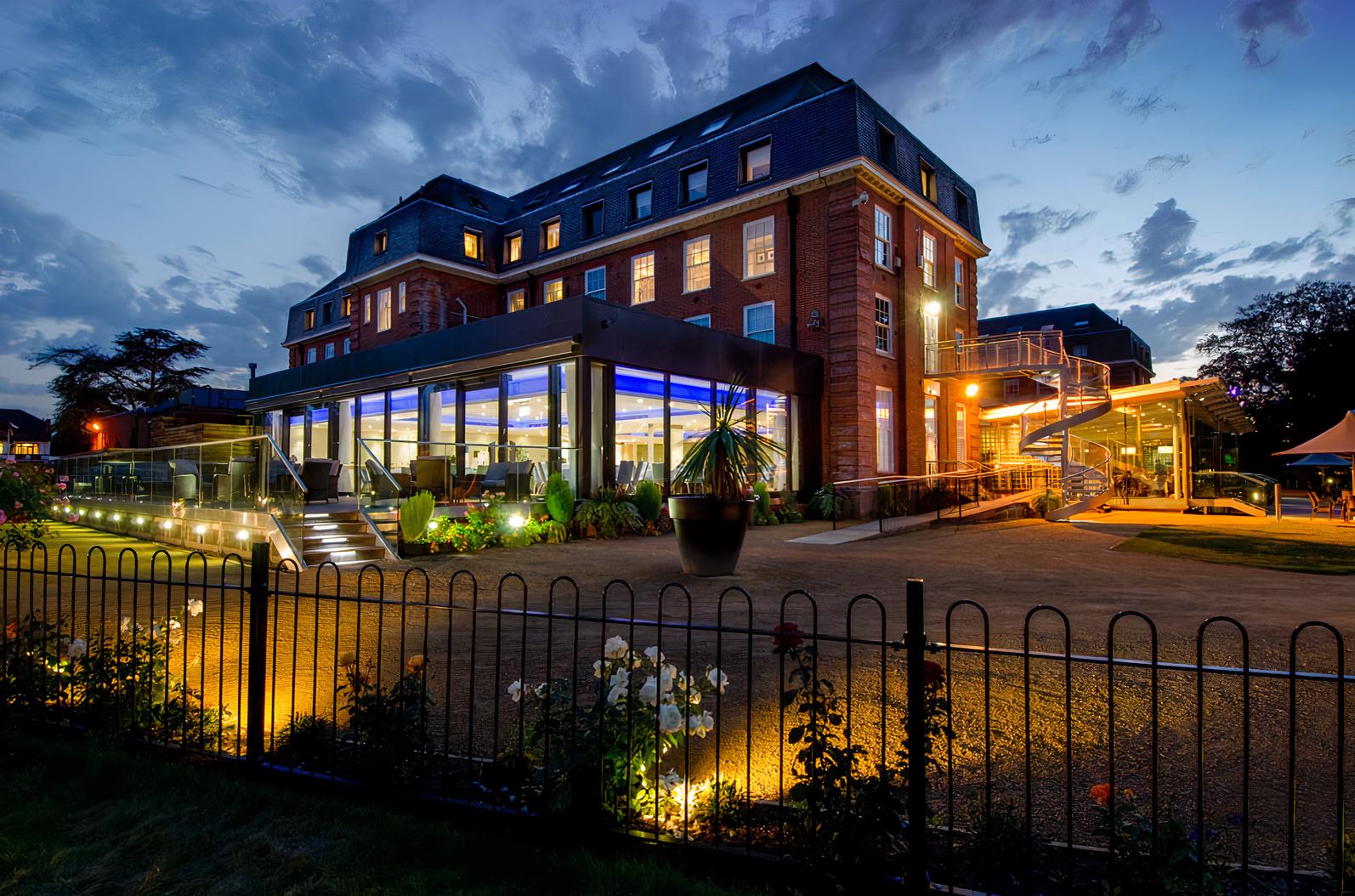 A hotel with illuminated exterior lighting, large windows, and landscaped gardens at The Lensbury.