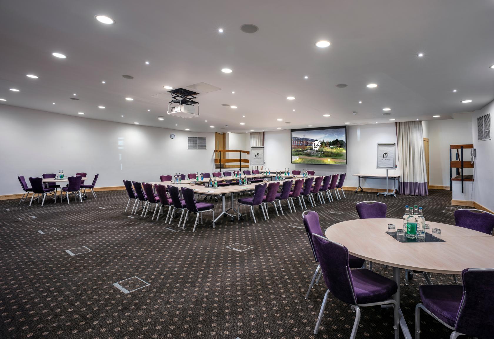 An auditorium with a large projection screen, round and rectangular tables, and purple chairs at The Lensbury Beaumont Room.
