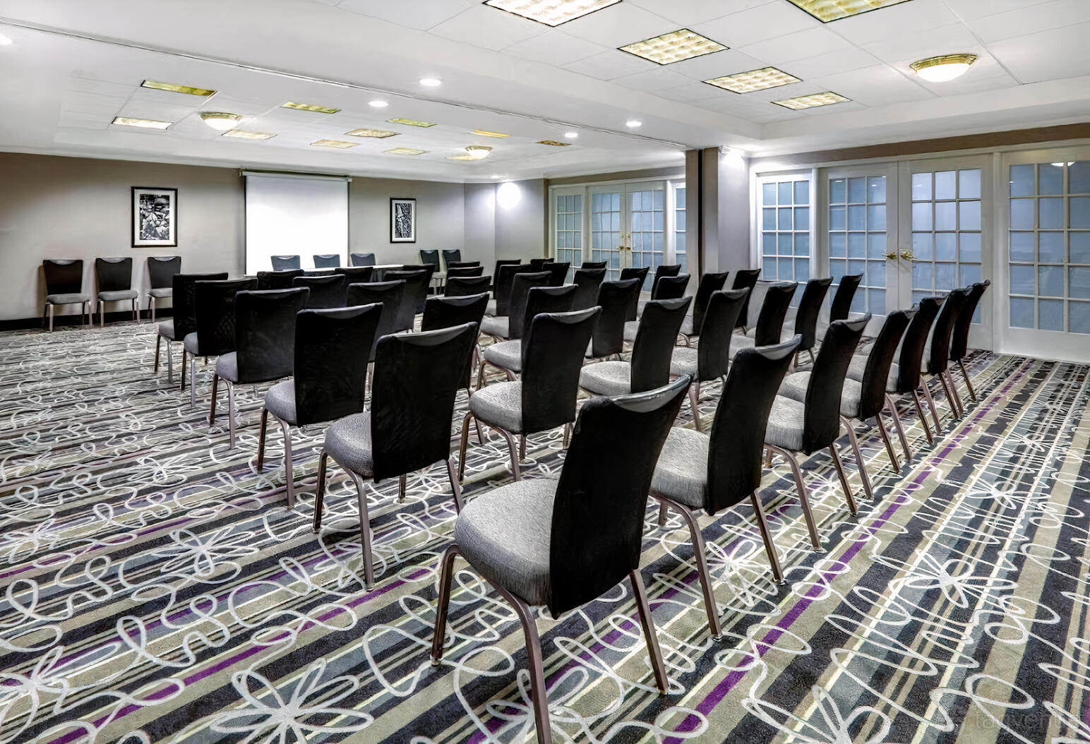 A meeting room with rows of black chairs, patterned carpet, and floor-to-ceiling windows at La Quinta Inn & Suites by Wyndham Dallas.