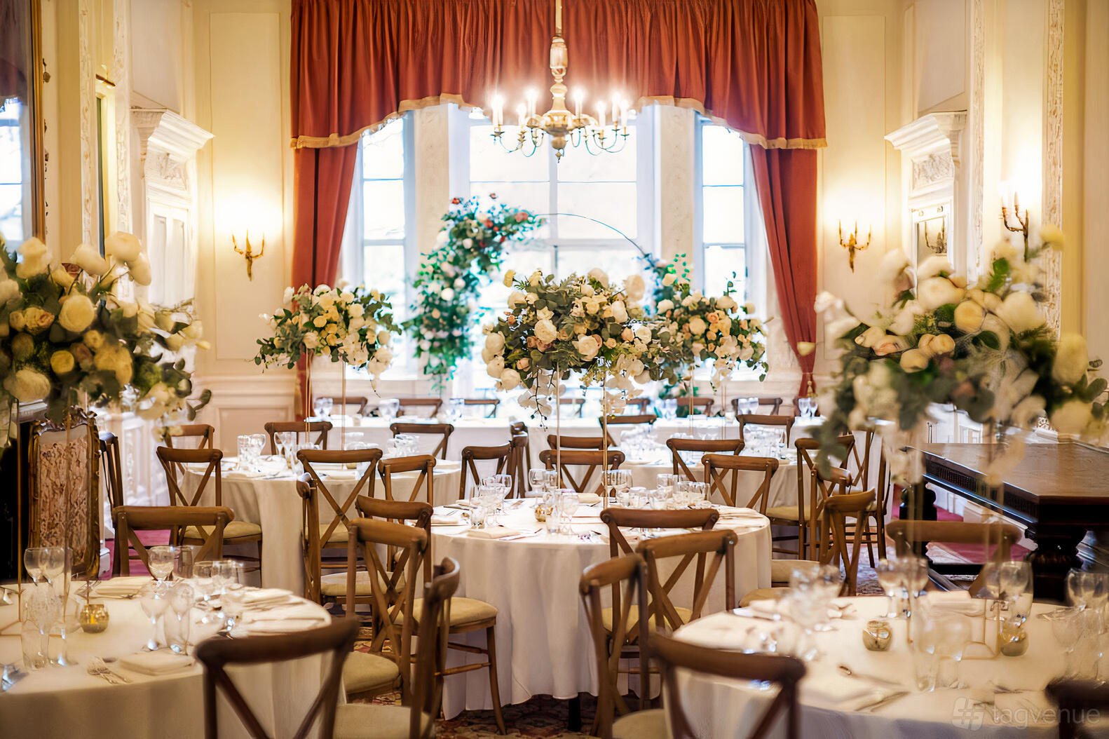 A function room with round tables, tall floral centerpieces, chandeliers, and large windows with red curtains at Six Park Place.