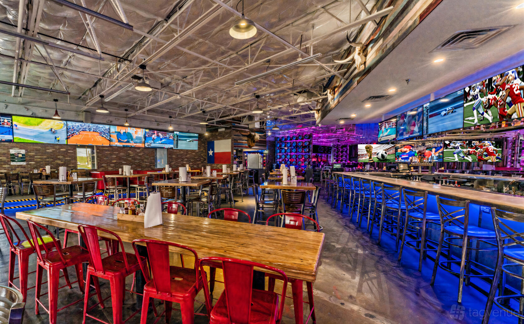 A bar with high ceilings, colorful metal chairs, long wooden tables, and multiple large TV screens at Longhorn Icehouse.