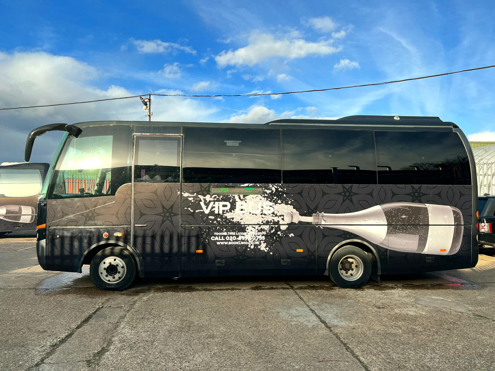 A black party bus with champagne bottle graphics parked on a concrete lot at Party Bus London.