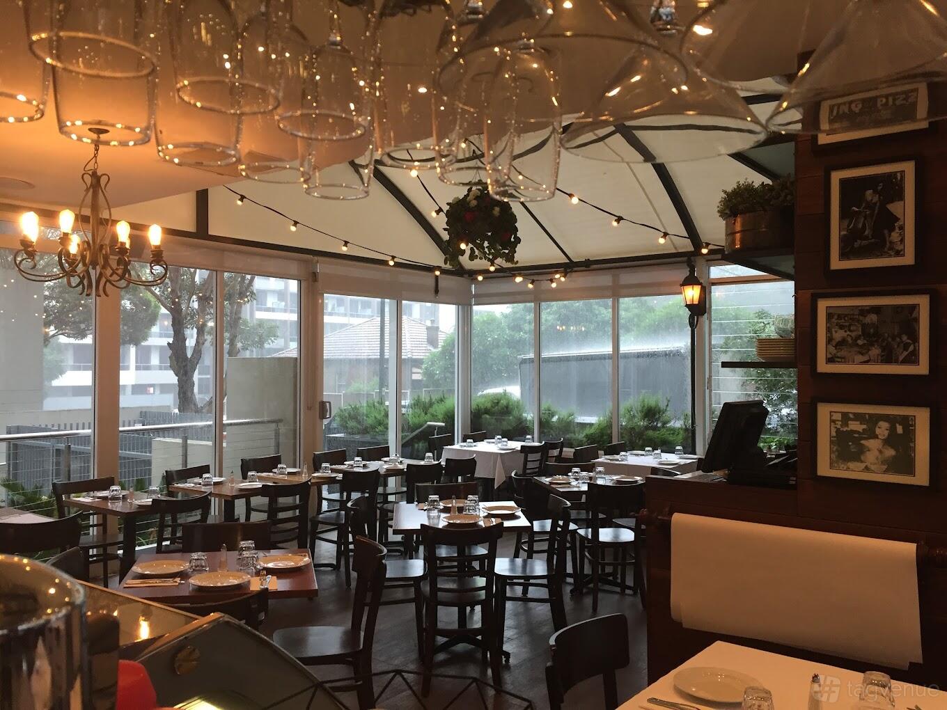 A restaurant with floor-to-ceiling windows, string lights, wooden tables, and hanging glassware at Andiamo Trattoria Rhodes.